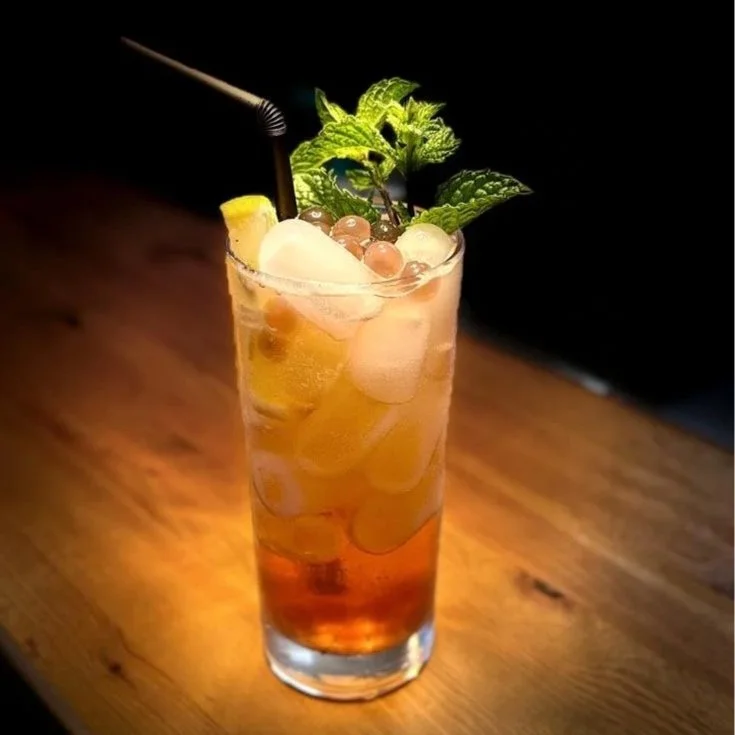 A tall glass of iced tea garnished with lemon slices, mint leaves, and tapioca pearls, with a black straw, placed on a wooden surface in a dimly lit environment.