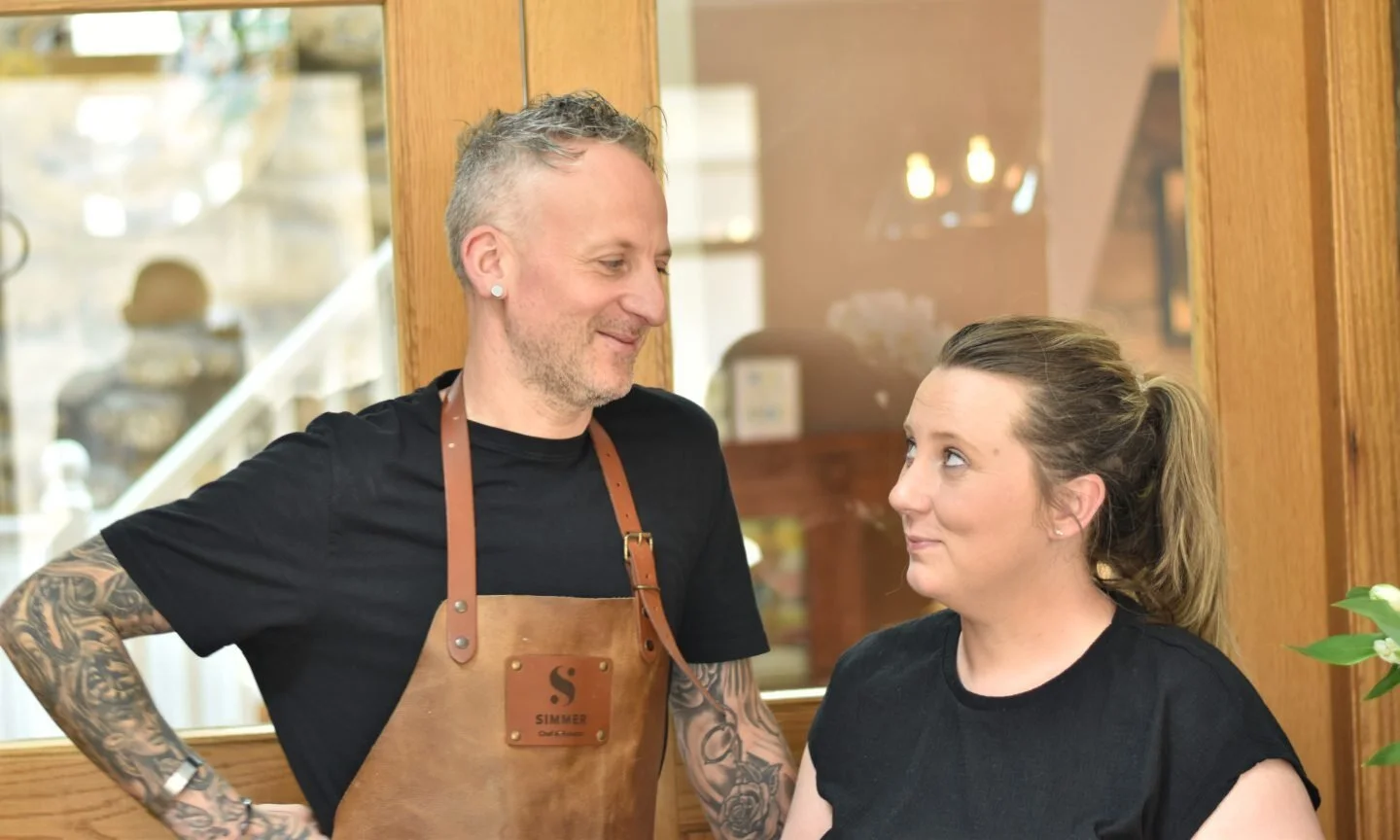 A man with tattoos and a woman with a ponytail standing indoors, looking at each other and smiling.