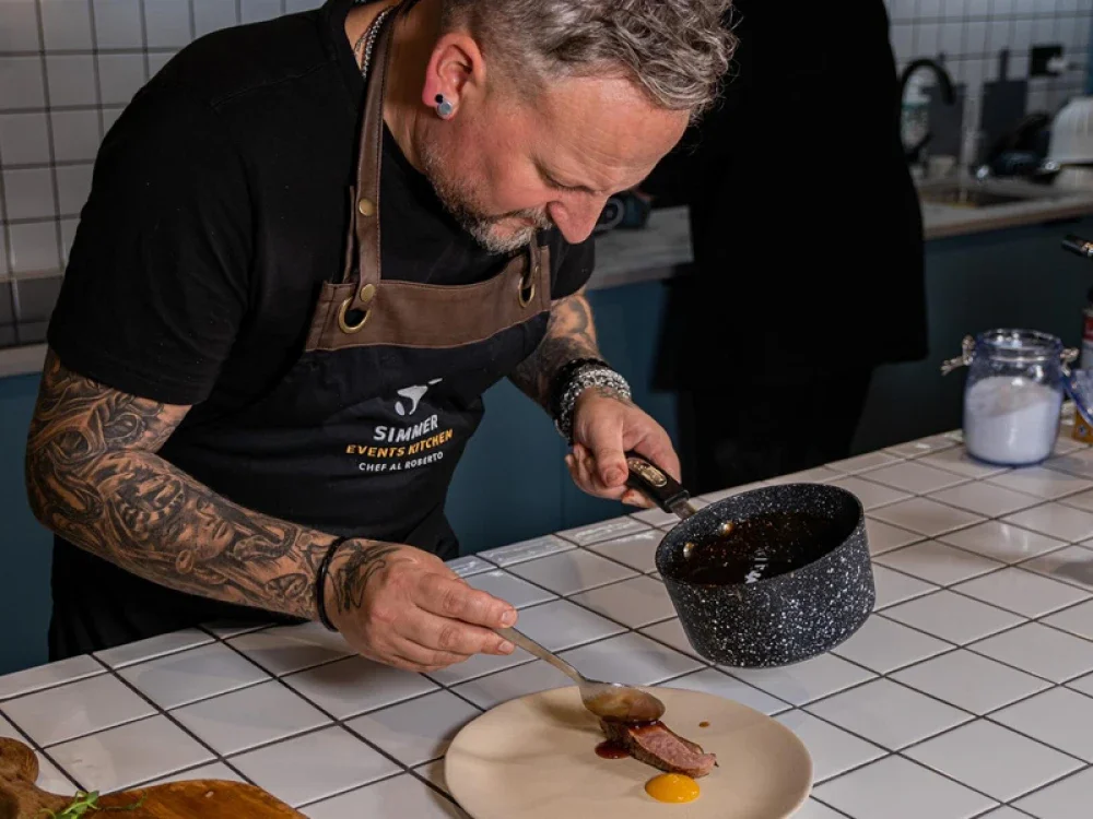 A man with tattoos and earrings, wearing a black shirt and apron, is sauce the plate with sauce from a pan in a kitchen with tiled counters.