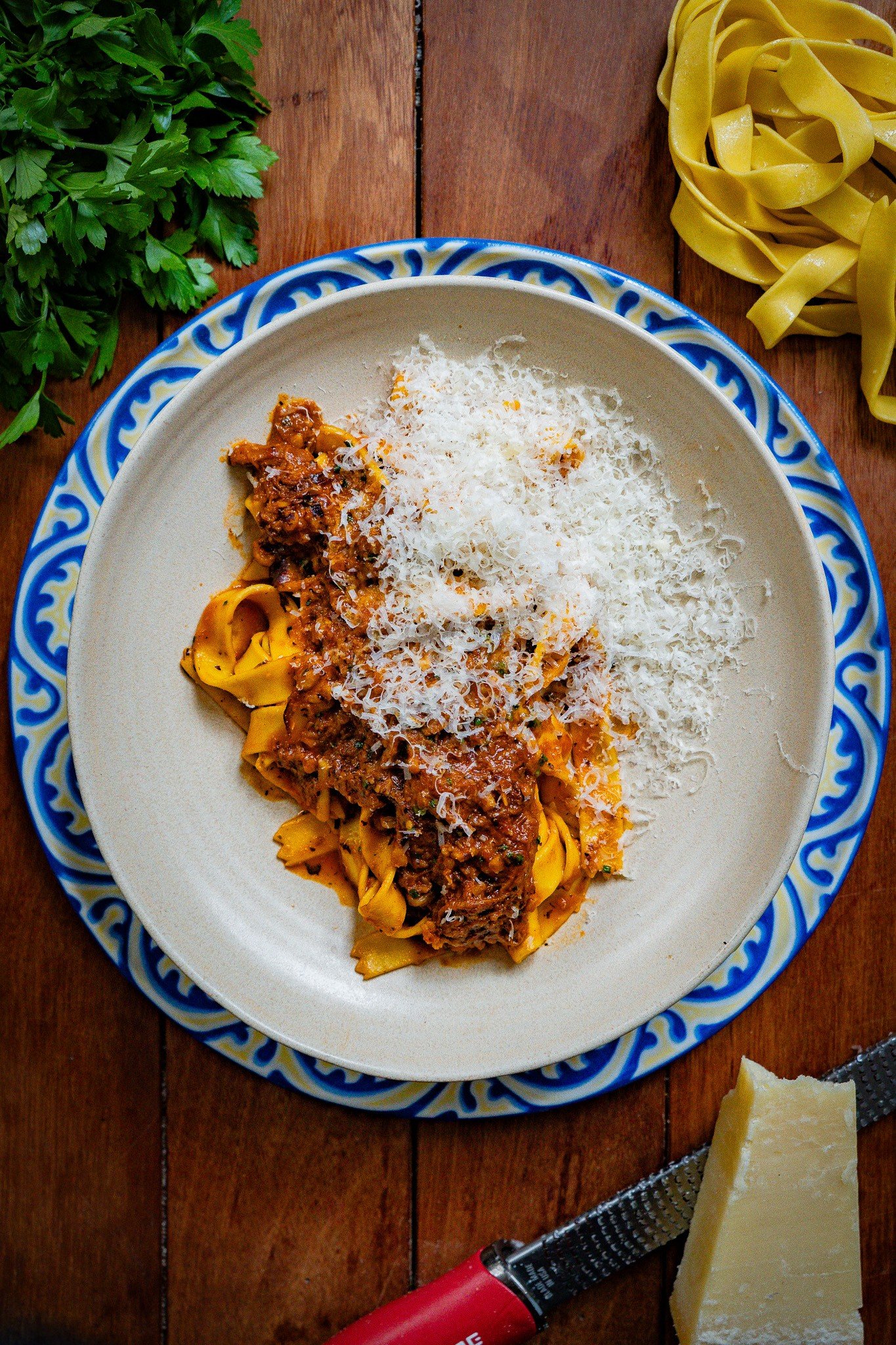 Plate of cooked pasta with meat sauce and grated cheese, fresh parsley on side, block of Parmesan cheese, and a pizza cutter on a wooden table.