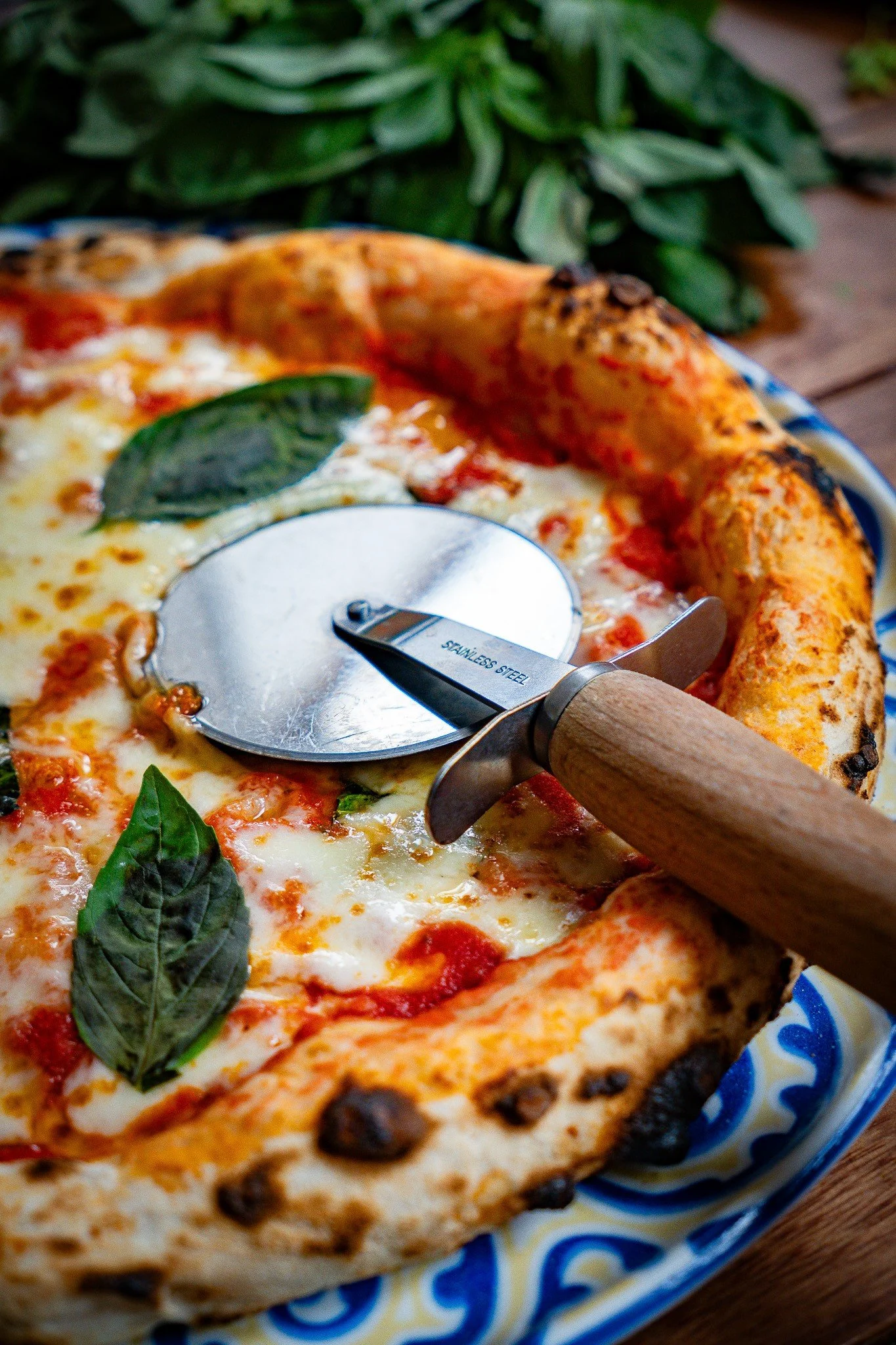 Close-up of a pizza with basil leaves, melted cheese, and toppings, with a pizza cutter on the plate.