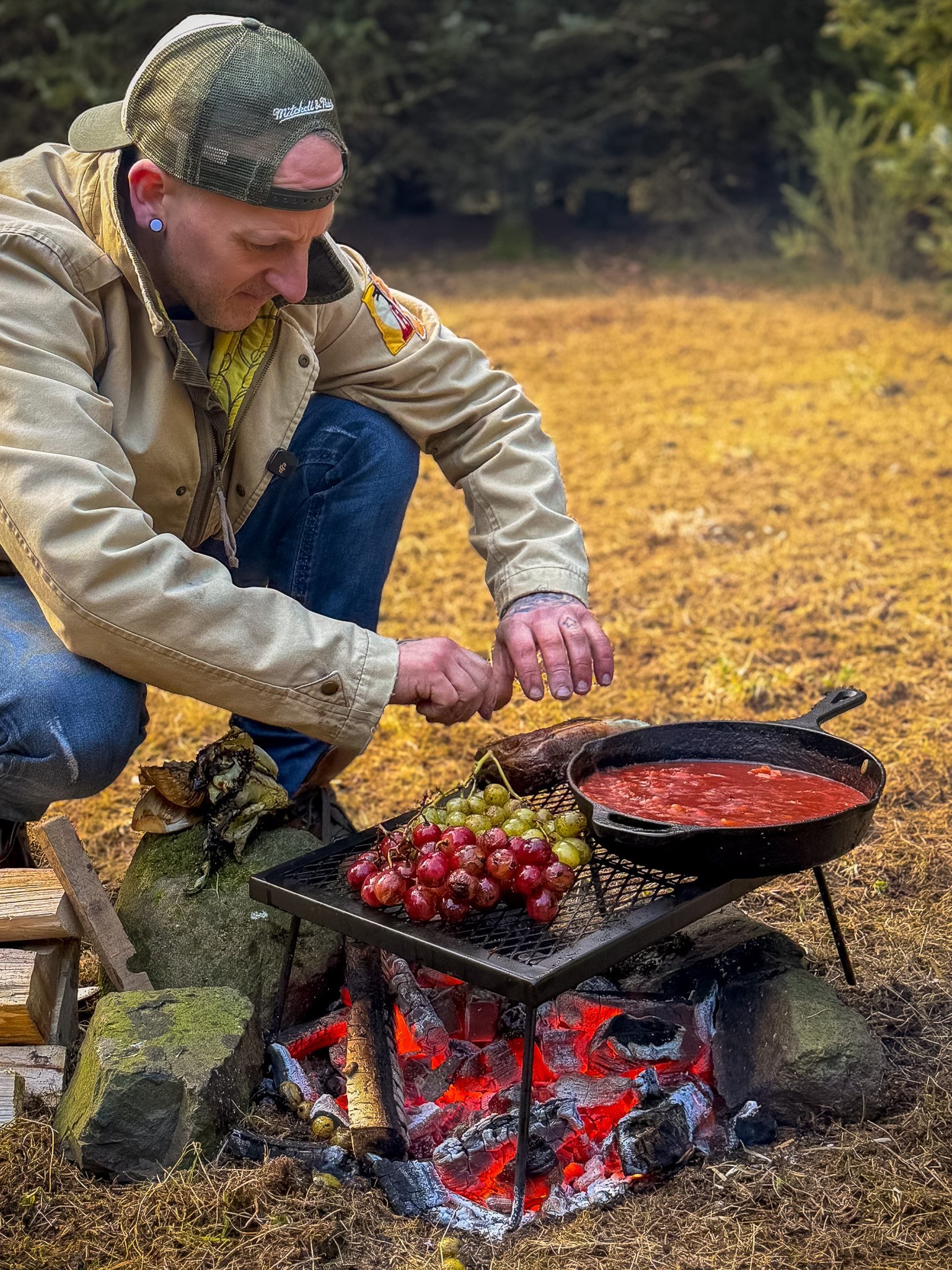 A man wearing a tan jacket, blue jeans, a camouflage cap, and earrings is crouching beside a campfire. The campfire has glowing coals and is cooking or heating a cast iron skillet filled with red liquid, possibly soup or sauce. A tray of grapes is on