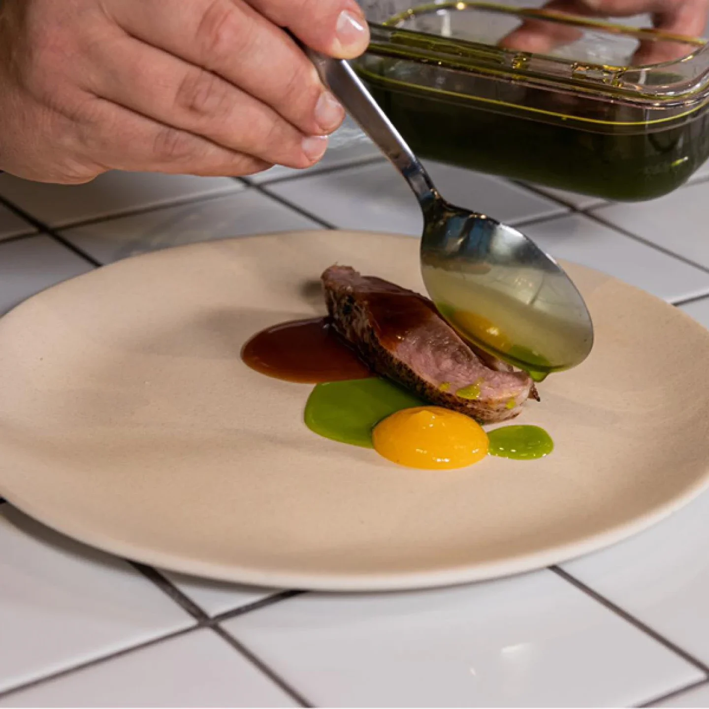 Chef drizzling sauce over a sliced medium-rare steak on a beige plate, accompanied by colorful sauces, on a white tiled kitchen counter.