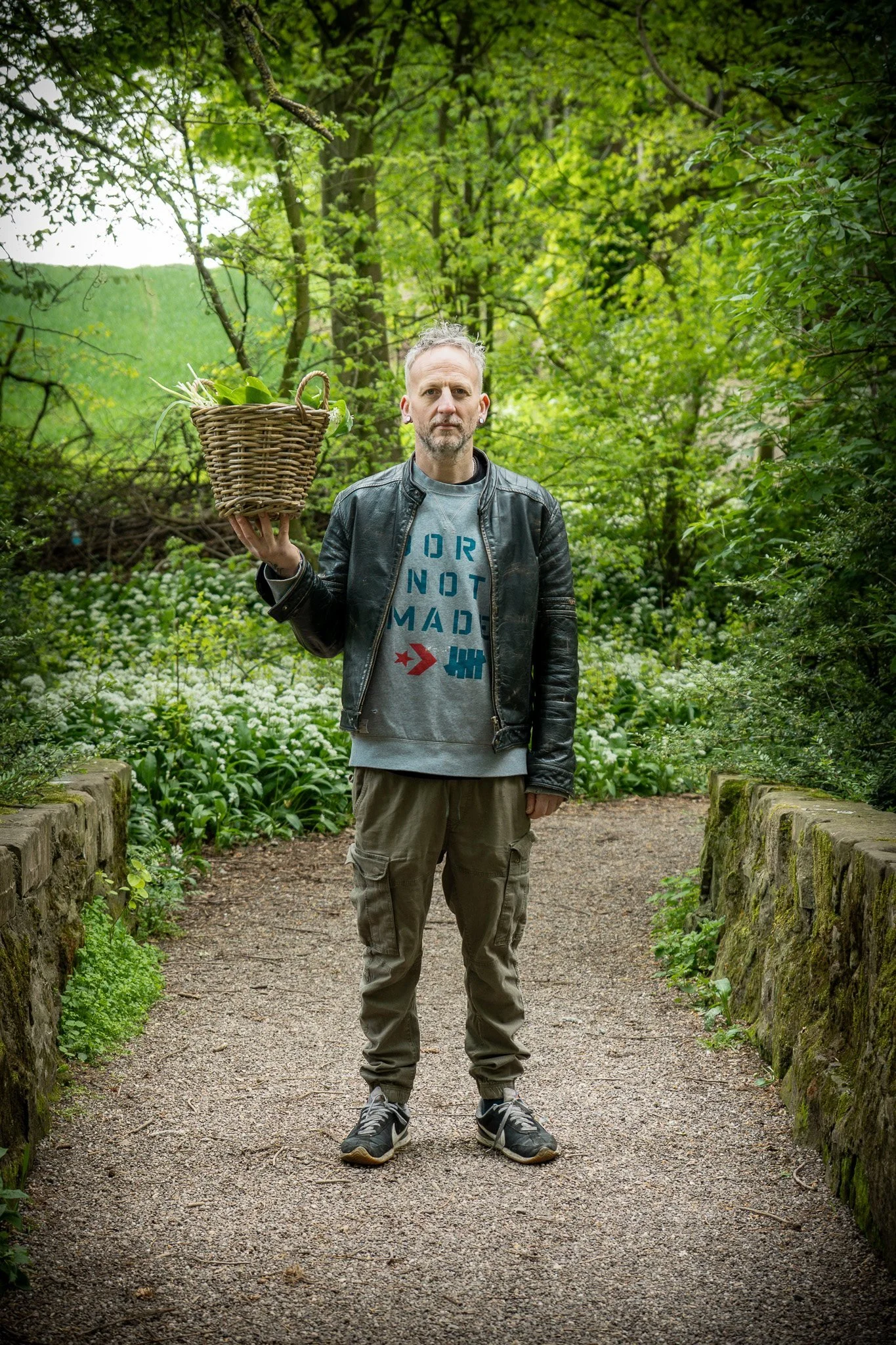 A man standing on a dirt path in a lush green forest, holding a wicker basket filled with leafy greens and herbs.
