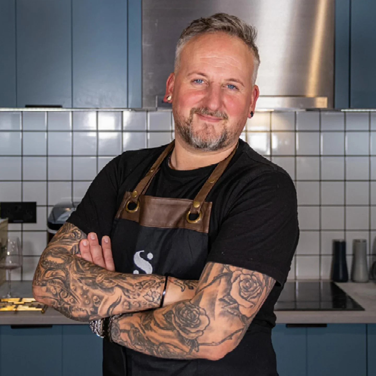 A man with short gray hair, a beard, tattoos on his arms, and earrings, standing in a modern kitchen with blue cabinets and white tiled backsplash, smiling with his arms crossed wearing a black t-shirt and a brown apron.