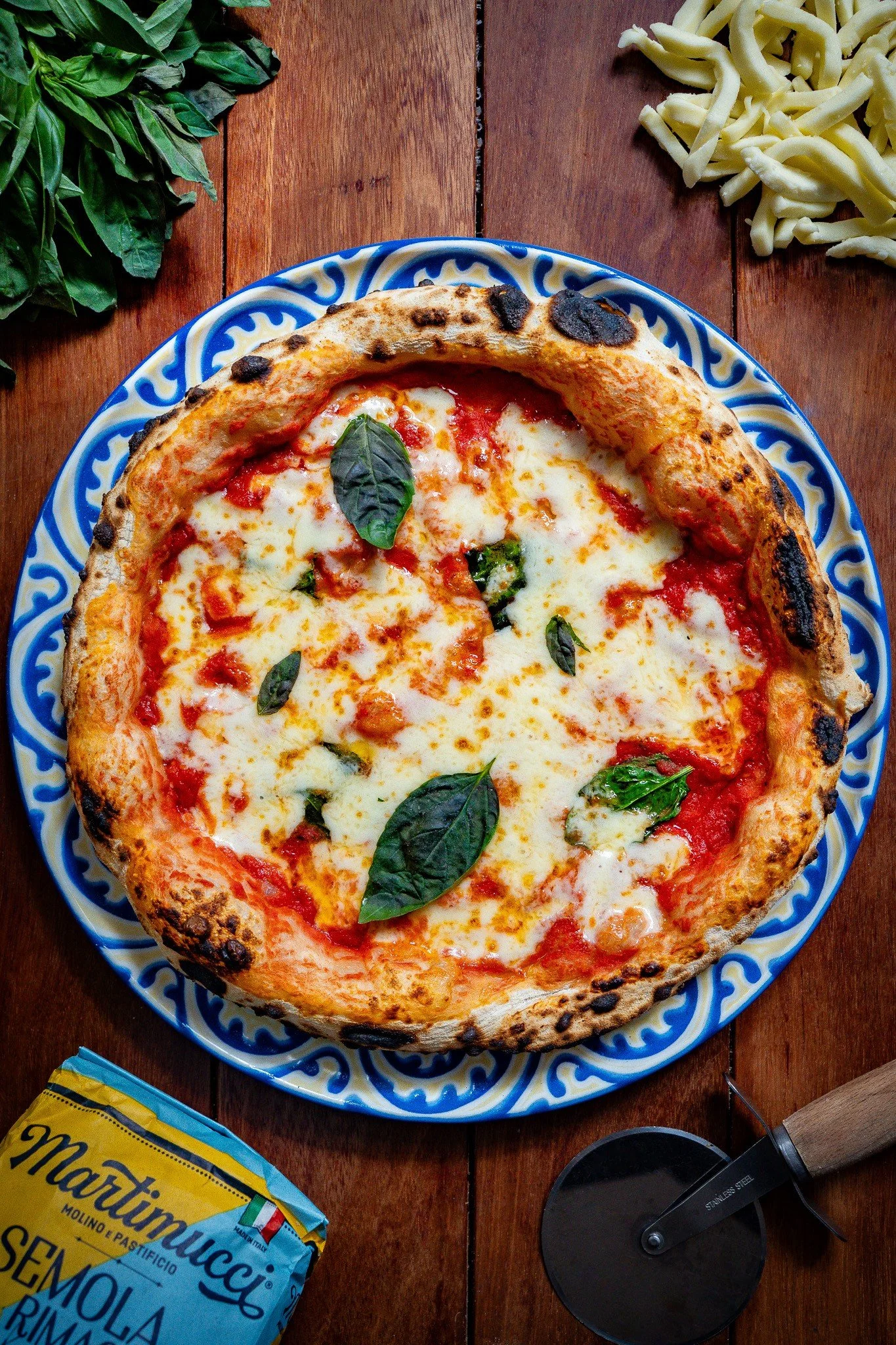 A margherita pizza with cheese, tomato sauce, and basil leaves on a blue and white patterned plate on a wooden table. Surrounding elements include a bunch of basil leaves to the top left, shredded cheese to the top right, a yellow bag of flour to the