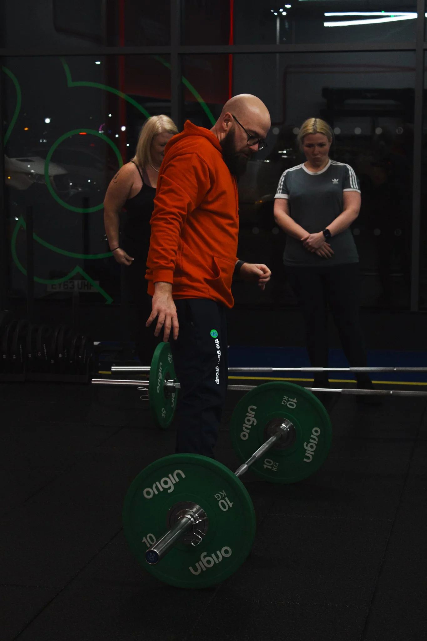 A man in an orange hoodie lifting a barbell with green weights in a gym, with two women observing behind him.