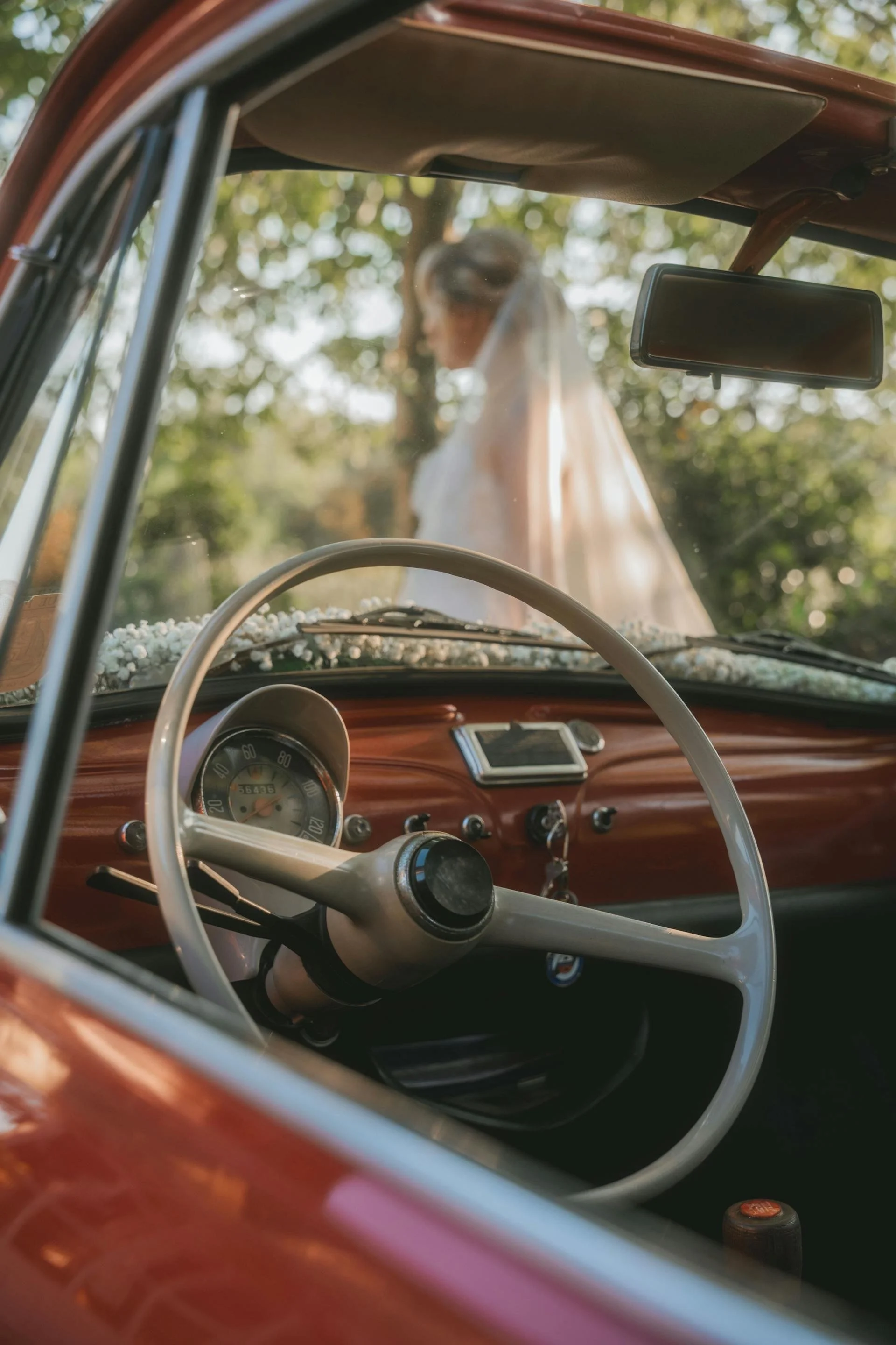View from inside a vintage car looking at a bride standing outside. The car has a wooden dashboard and a large steering wheel. The bride is blurred in the background, wearing a white dress and veil, with trees in the background.