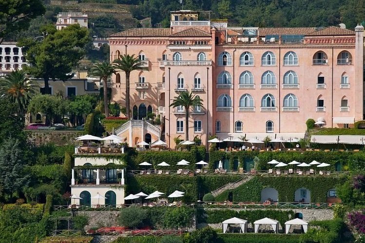 A large pink hotel building with multiple balconies, surrounded by lush greenery, palm trees, and outdoor patio areas with tables and umbrellas on a hillside.