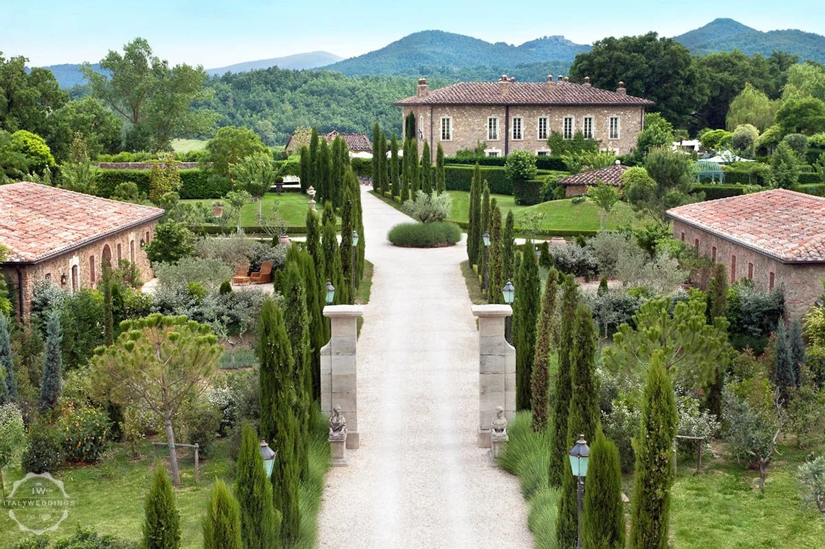 A scenic view of a large estate with a gravel pathway lined with tall cypress trees, leading to a main stone house with red-tiled roof, surrounded by lush green gardens and rolling hills in the background.