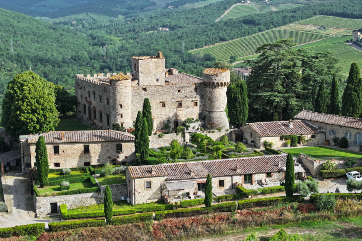 A medieval castle surrounded by lush greenery and smaller stone buildings, located in a scenic countryside with rolling hills and vineyards.