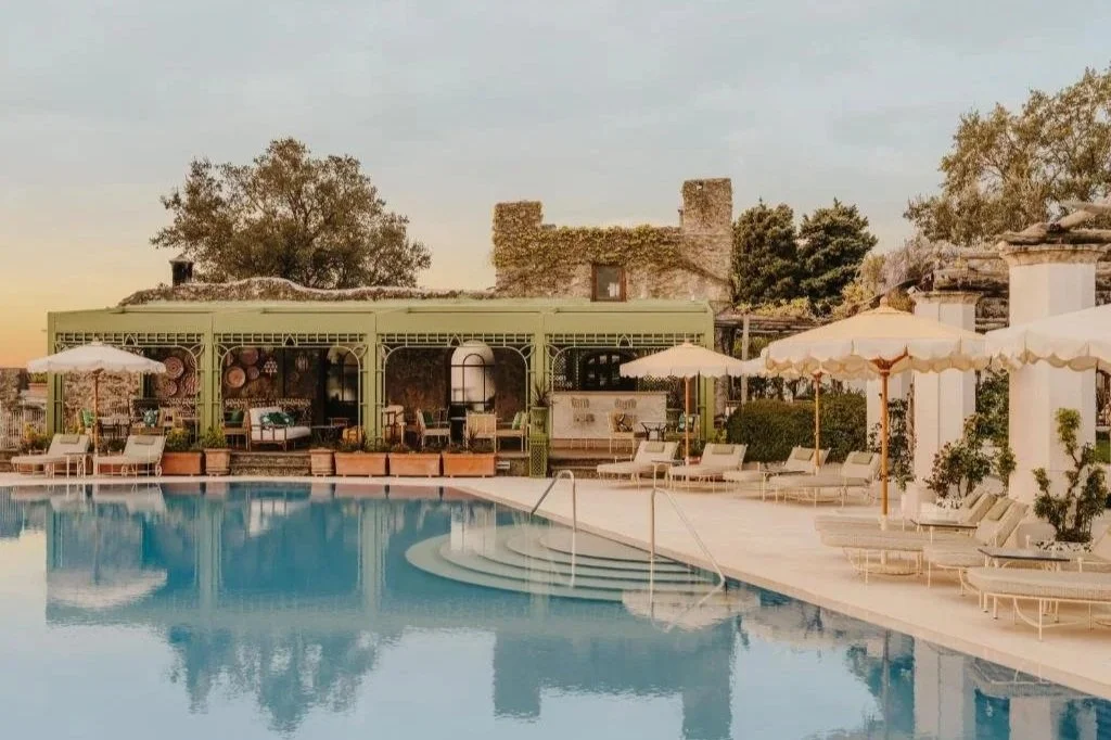 A luxurious outdoor swimming pool area with lounge chairs, umbrellas, and a green shaded cabana, with a rustic stone building and trees in the background during sunset.