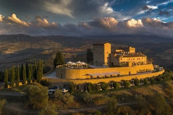 Castle-like building on a hill with a sunset sky, surrounded by cypress trees and parked cars.