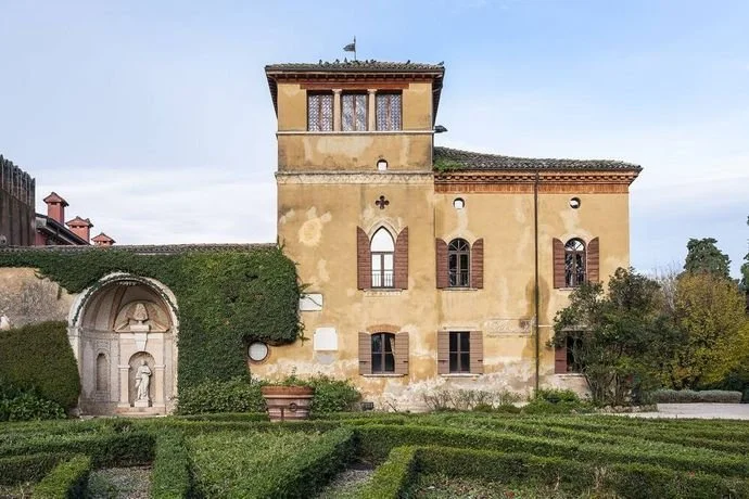 A historic European building with a tower, arched windows, and a stone fountain with a carved figure, surrounded by a well-maintained garden with hedges.