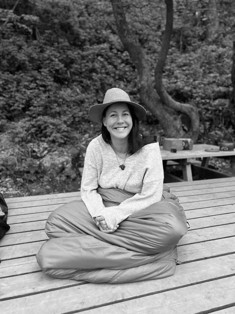 Frau mit schwarzen Haaren und braunem Hut, sitzend auf Holzplattform im Grünen, lächelnd.