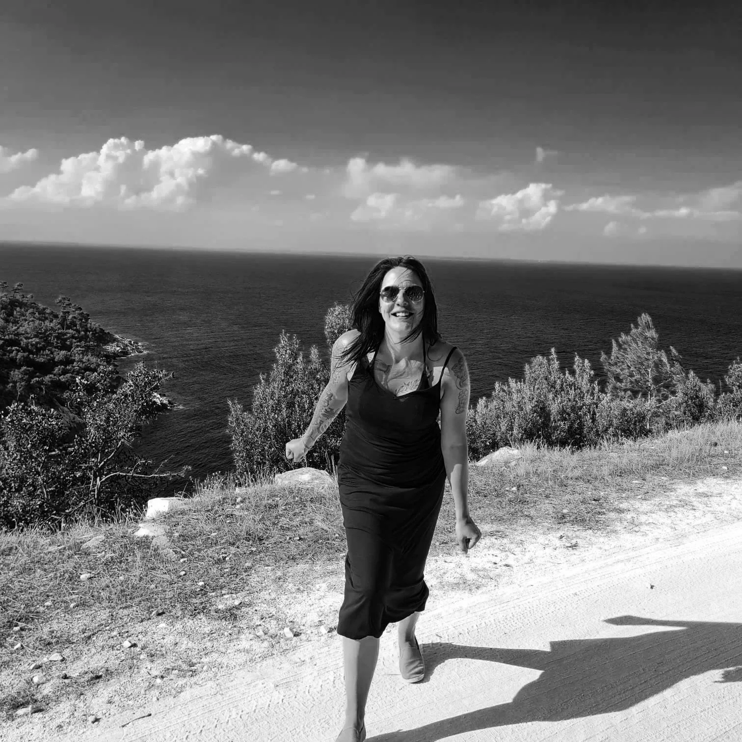 Frau in schwarzem Kleid lachend an der Küste mit Blick auf das Meer, Sonnenschein, blauer Himmel.