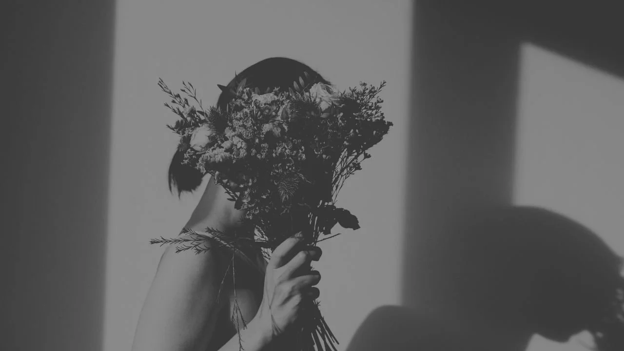Schwarzes und weißes Foto einer Person, die ein Blumenbouquet vor das Gesicht hält, sodass nur die Hand und das Bouquet sichtbar sind, mit Schatten an der Wand im Hintergrund.