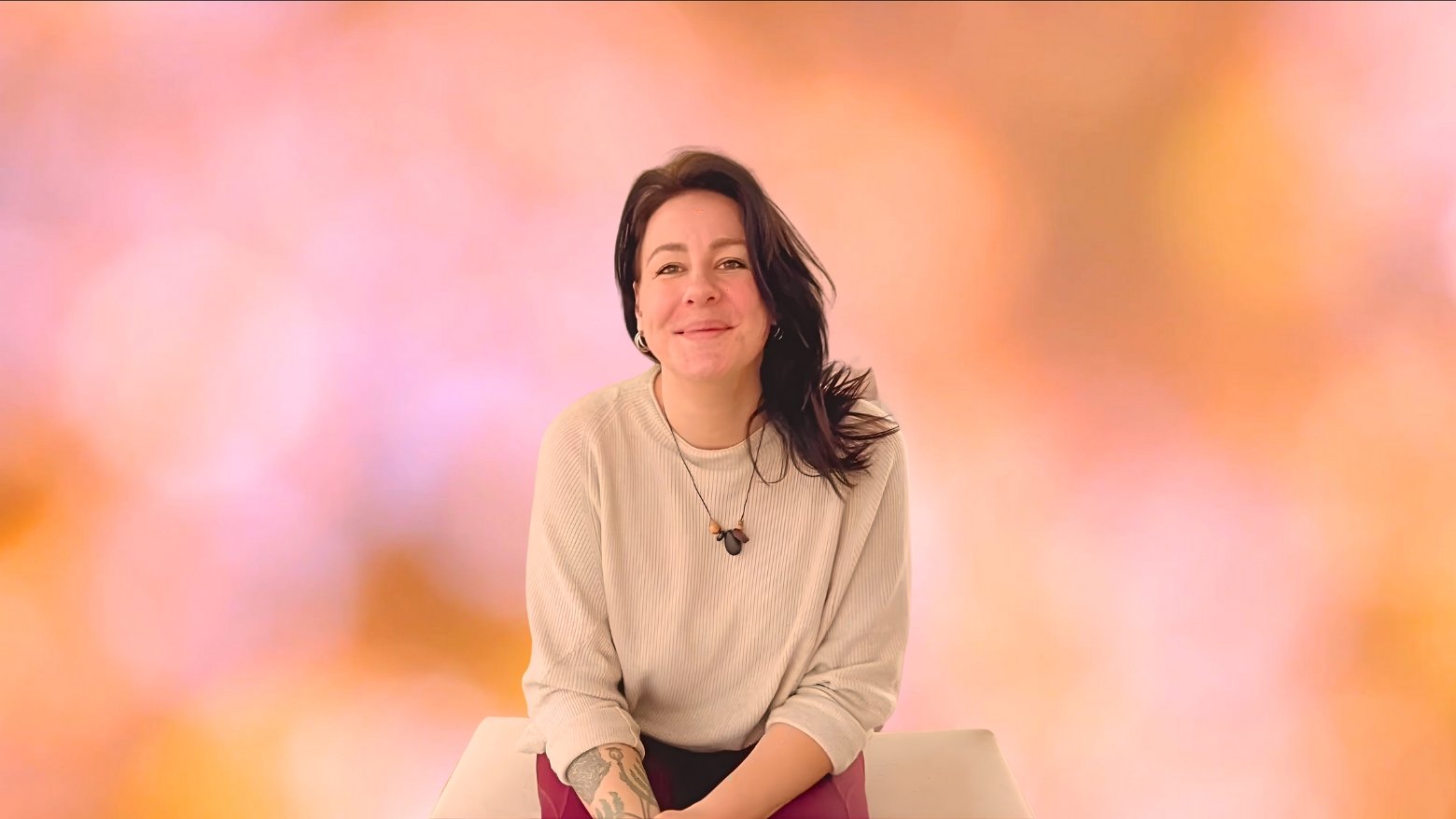 Junge Frau mit dunklen Haaren, lockeren Kleidung und Tattoos, sitzt auf einem weißen Hocker vor einem bunten, unscharfen Hintergrund, lächelt.