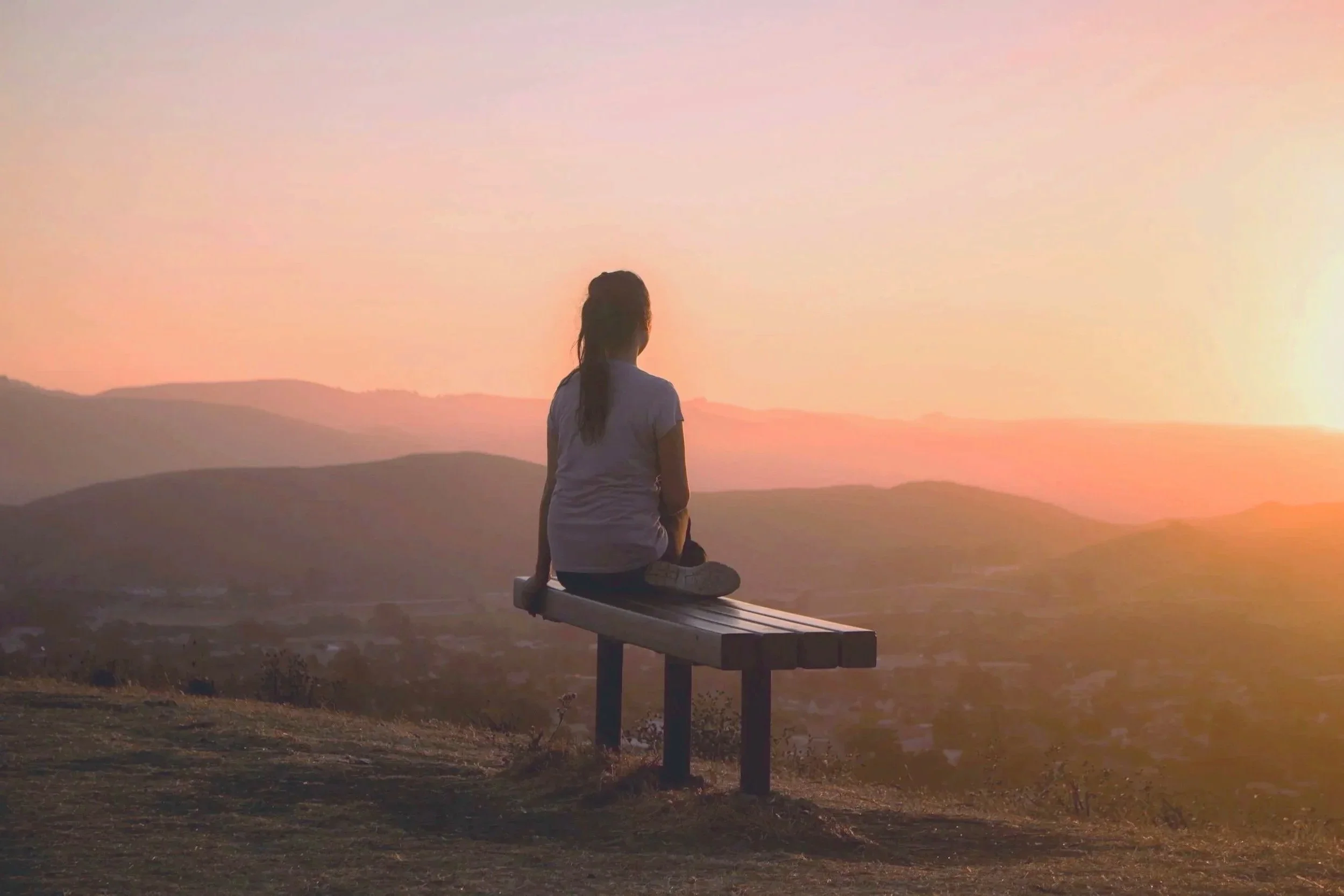 Ein Mädchen sitzt auf einer Bank und blickt auf eine Sonnenuntergangslandschaft mit Hügeln und einem weiten Himmel im Hintergrund.