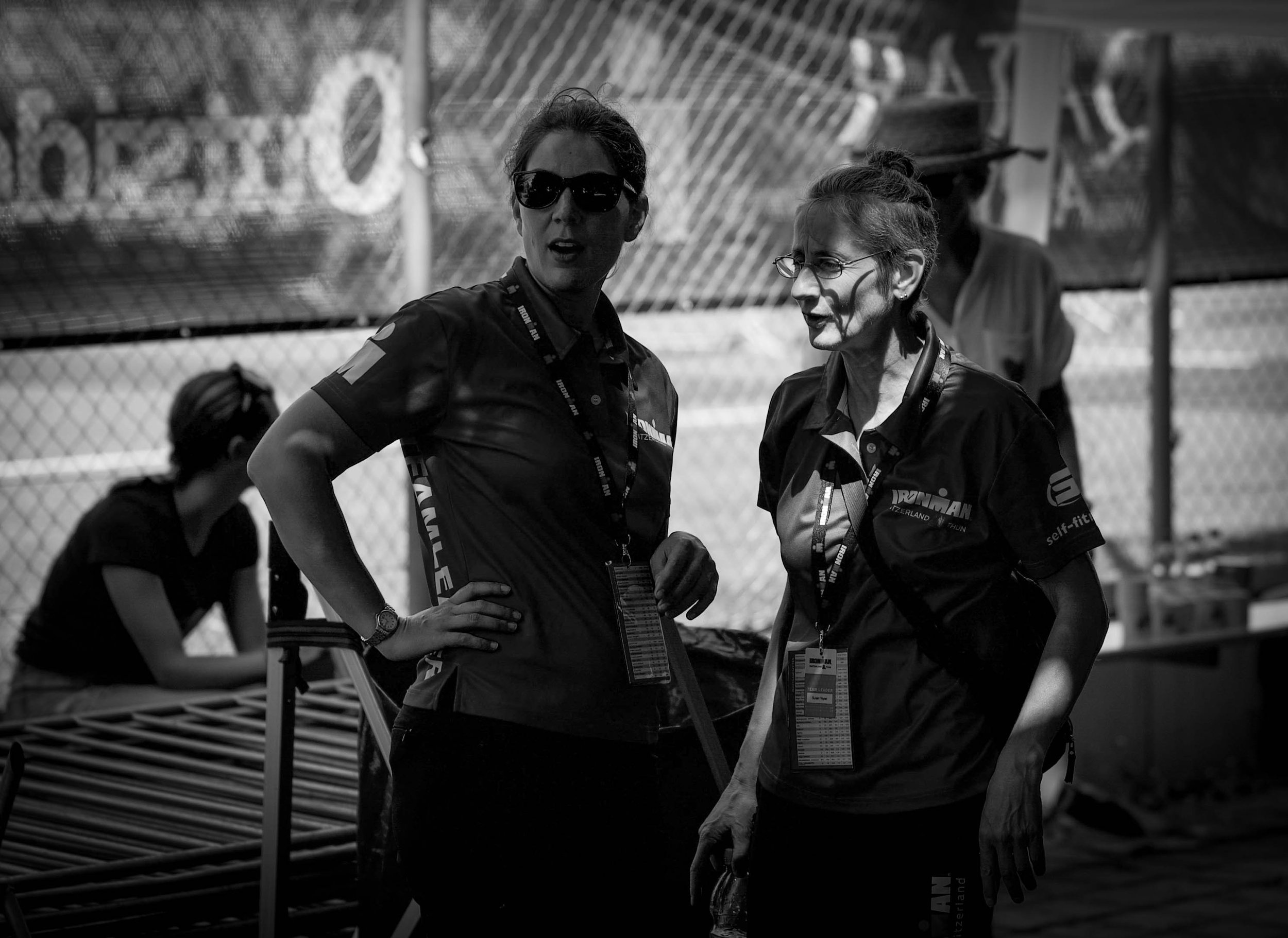 Zwei Frauen in Teamuniformen sprechen während eines Sonnentags an einem Sportevent, im Hintergrund eine weitere Person und ein Zaun.