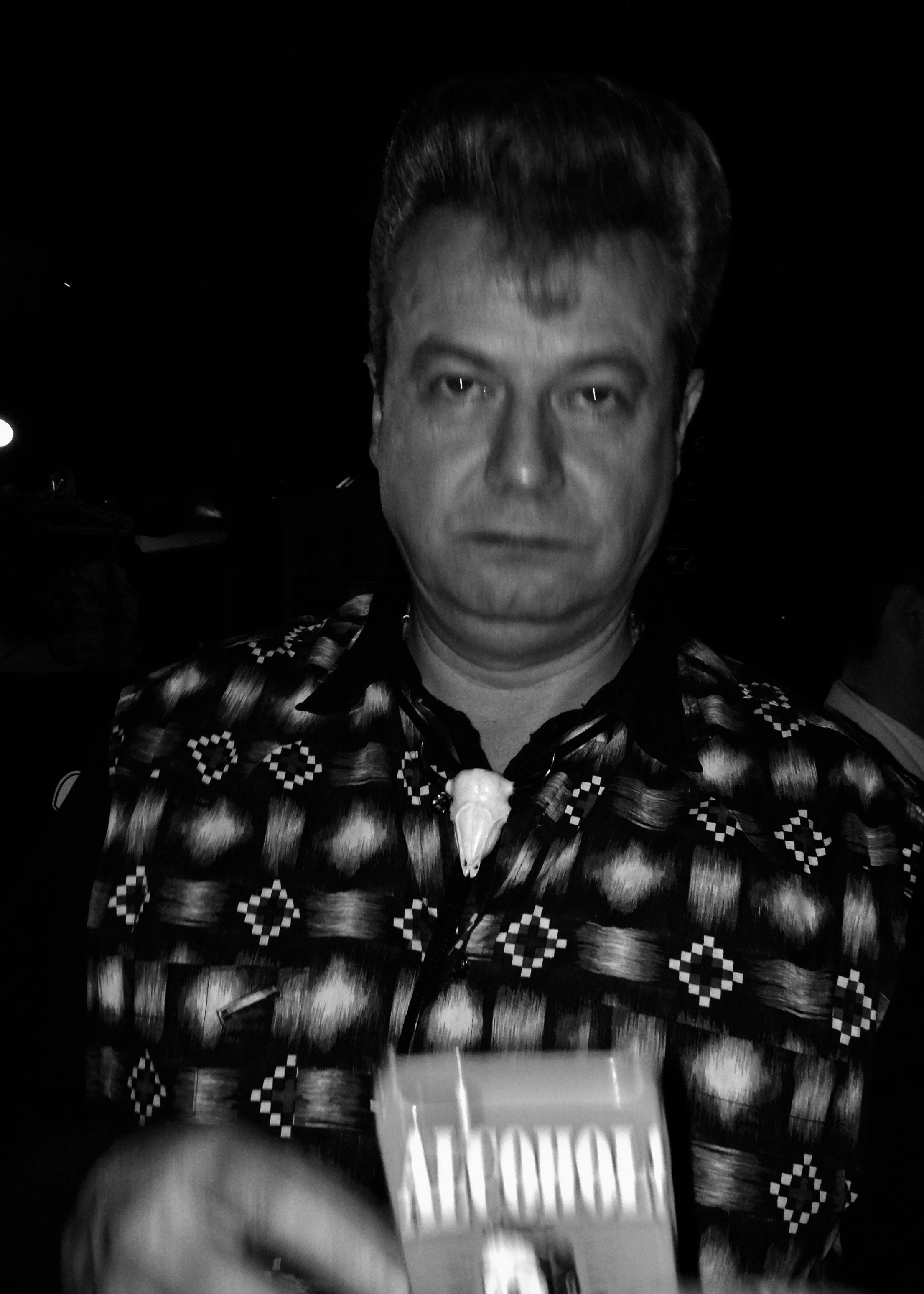 Schwarz-weiß-Fotografie eines Mannes mit vollem, dunklem, hochgestecktem Haar, trägt ein Hemd mit geometrischem Muster und eine Halskette mit einem Tierkopf-Anhänger, vor dunklem Hintergrund.