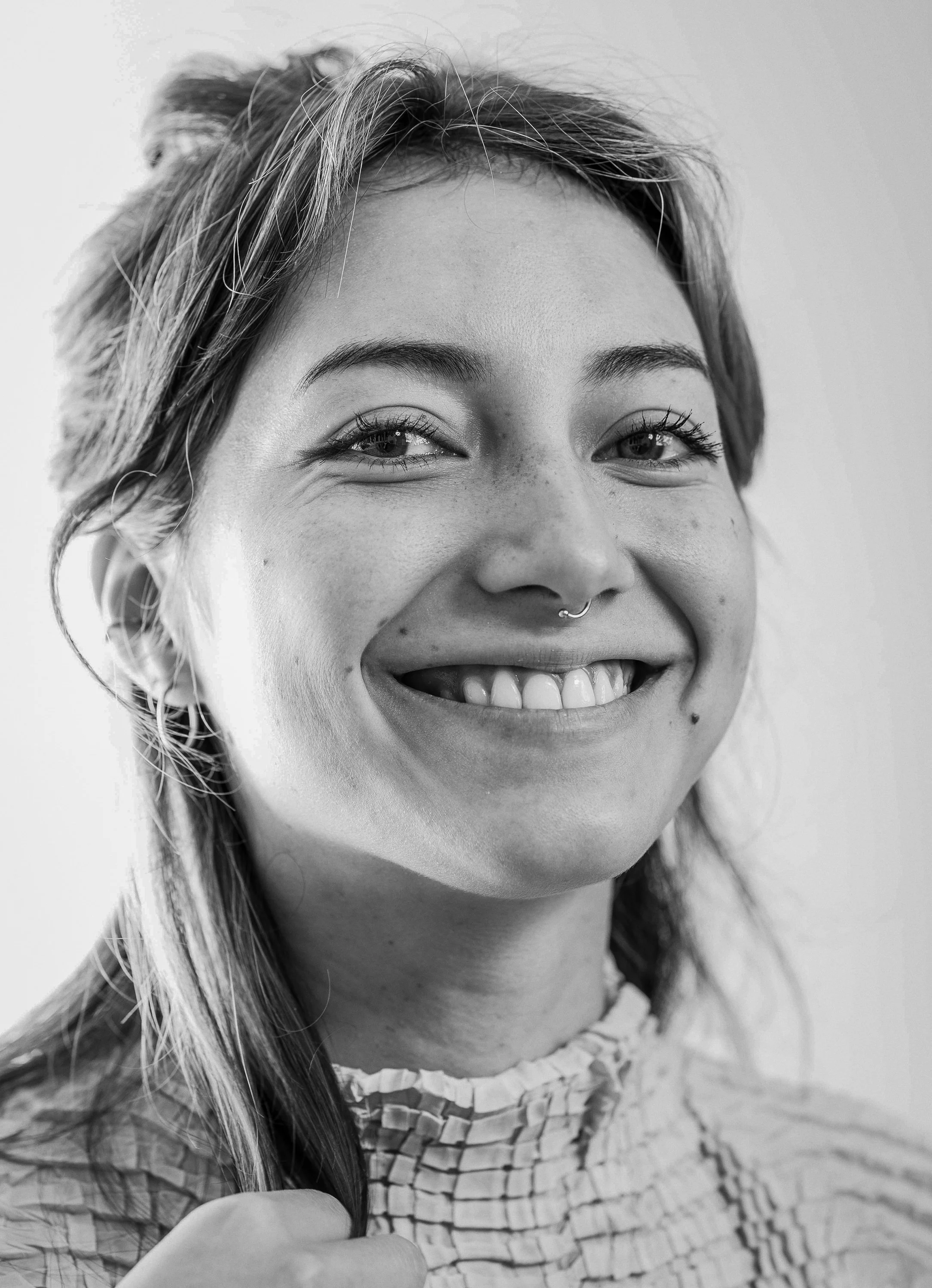 Schwarz-weiß Porträt einer lachenden jungen Frau mit einem Piercing an der Nase, lockigem Haar, Haut mit Sommersprossen, trägt ein geriptes Oberteil.