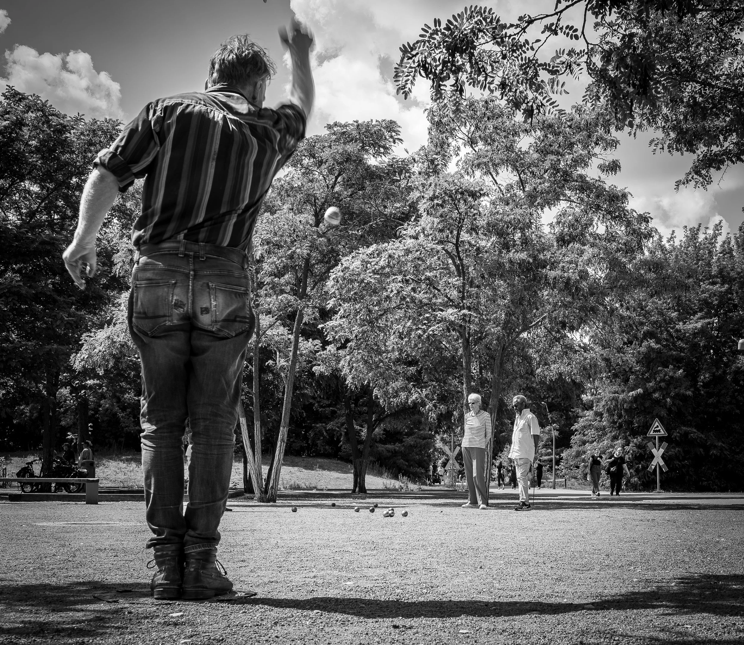 Menschen spielen Boule im Park, im Vordergrund ein Mann wirft eine Kugel, im Hintergrund drei weitere Personen und Bäume
