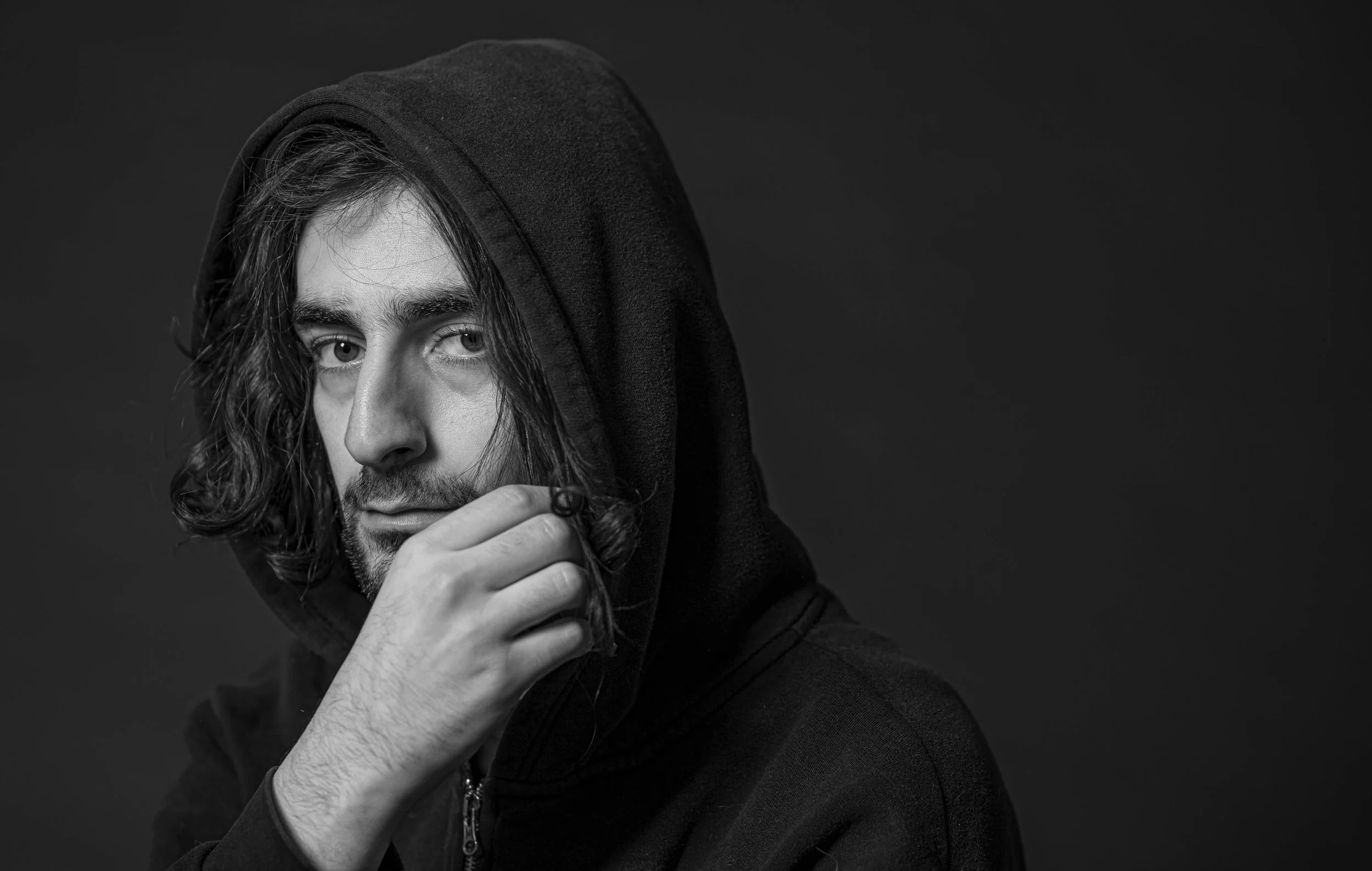 Schwarz-Weiß-Porträt eines jungen Mannes mit langen, lockigen Haaren, der einen schwarzen Hoodie trägt. Er berührt sein Kinn mit der Hand und schaut nachdenklich in die Kamera.