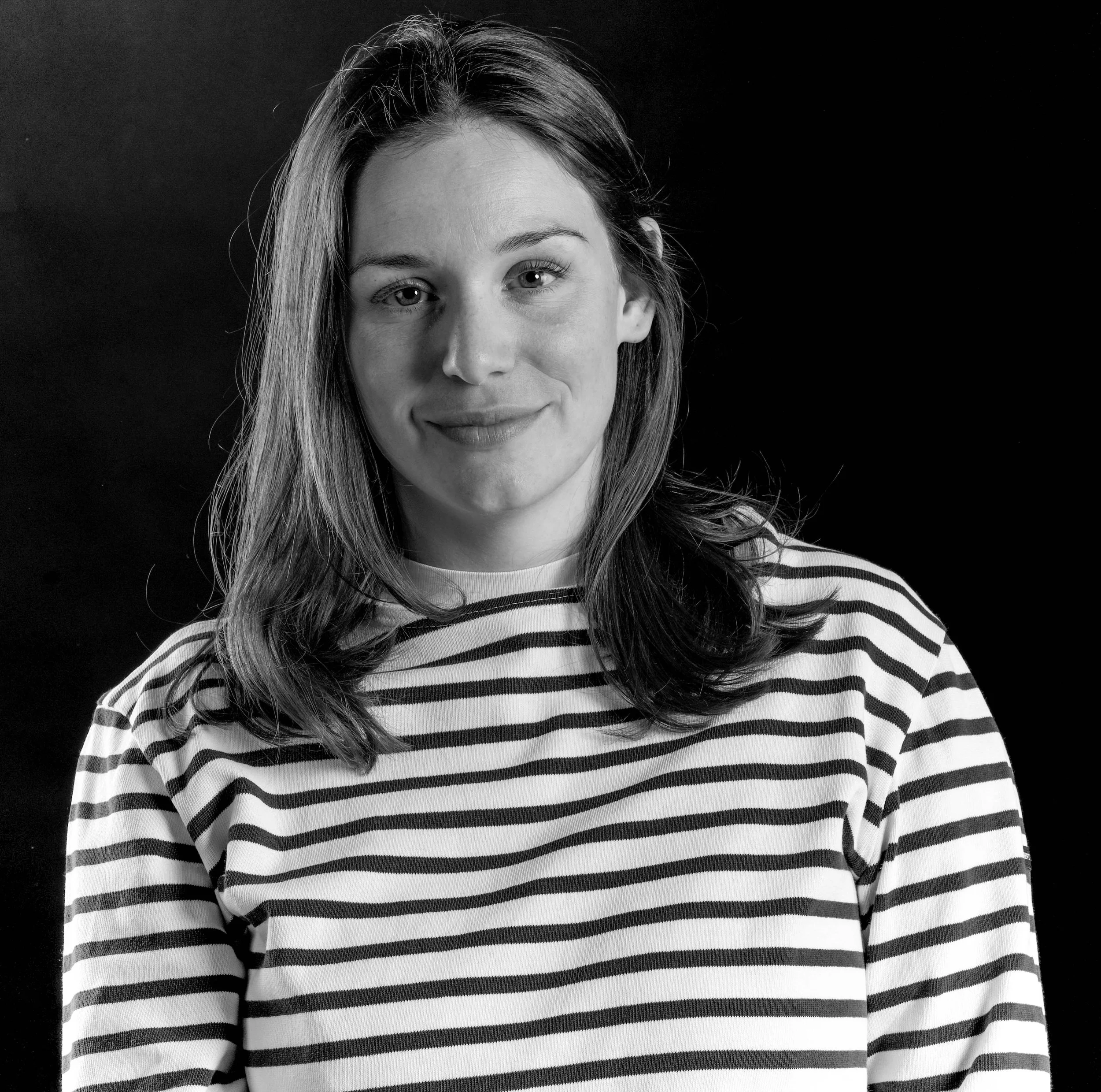 Schwarz-weiß Portrait einer lächelnden jungen Frau mit Fachwerk-gestreiftem Pullover vor dunklem Hintergrund.