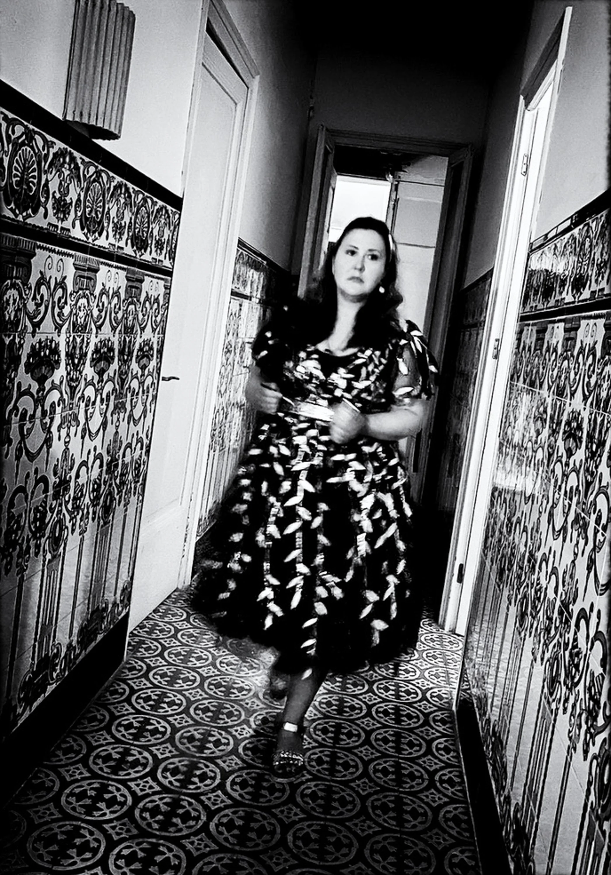 Schwarz-weiß-Foto einer Frau in einem eleganten Kleid, die in einem Flur steht, mit dekorativen Fliesen an den Wänden und einem offenen Tür im Hintergrund.