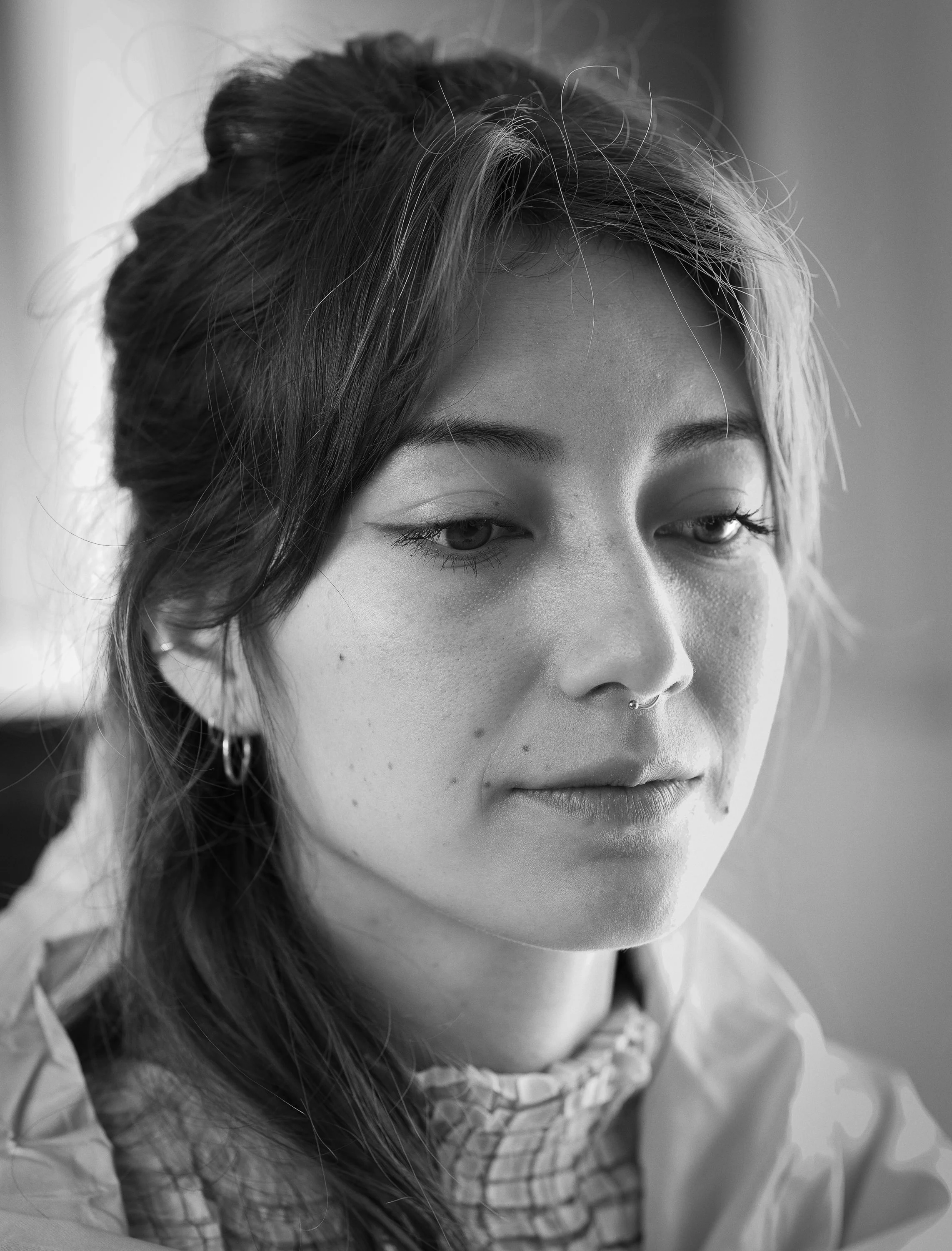 Schwarz-Weiß-Foto eines jungen Mädchens mit dunklen Haaren, Nase mit Piercing, und kleinen Ohrringen, das nachdenklich aussieht.