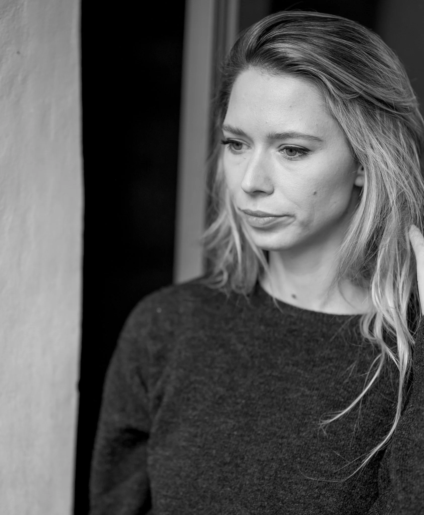 Schwarz-weiß-Foto einer jungen Frau mit langem, gewelltem Haar, die an einer Wand lehnte und nachdenklich blickt.