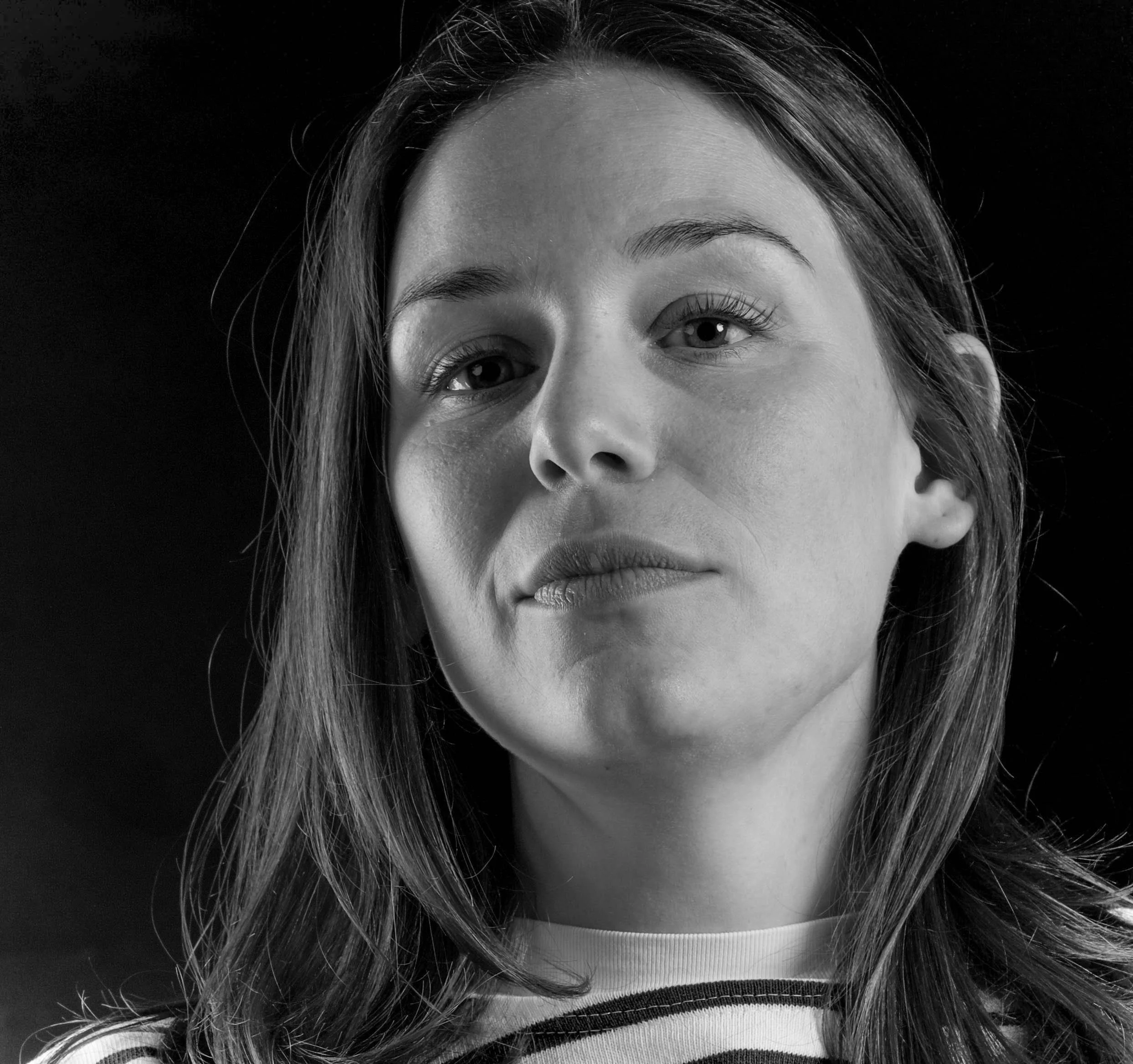 Schwarz-weiß Porträt einer jungen Frau mit mittellangem Haar, direkte Blick in die Kamera, neutraler Ausdruck, dunkler Hintergrund.