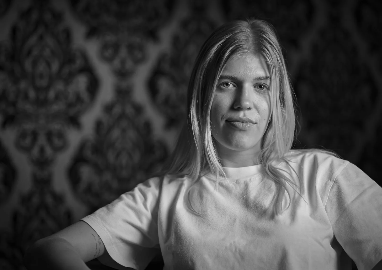 Schwarz-Weiß-Fotografie eines jungen Frau mit mittellangem Haar, die direkt in die Kamera schaut, lächelt leicht und entspannt wirkt, mit einem detaillierten, ornamentalen Muster im Hintergrund.