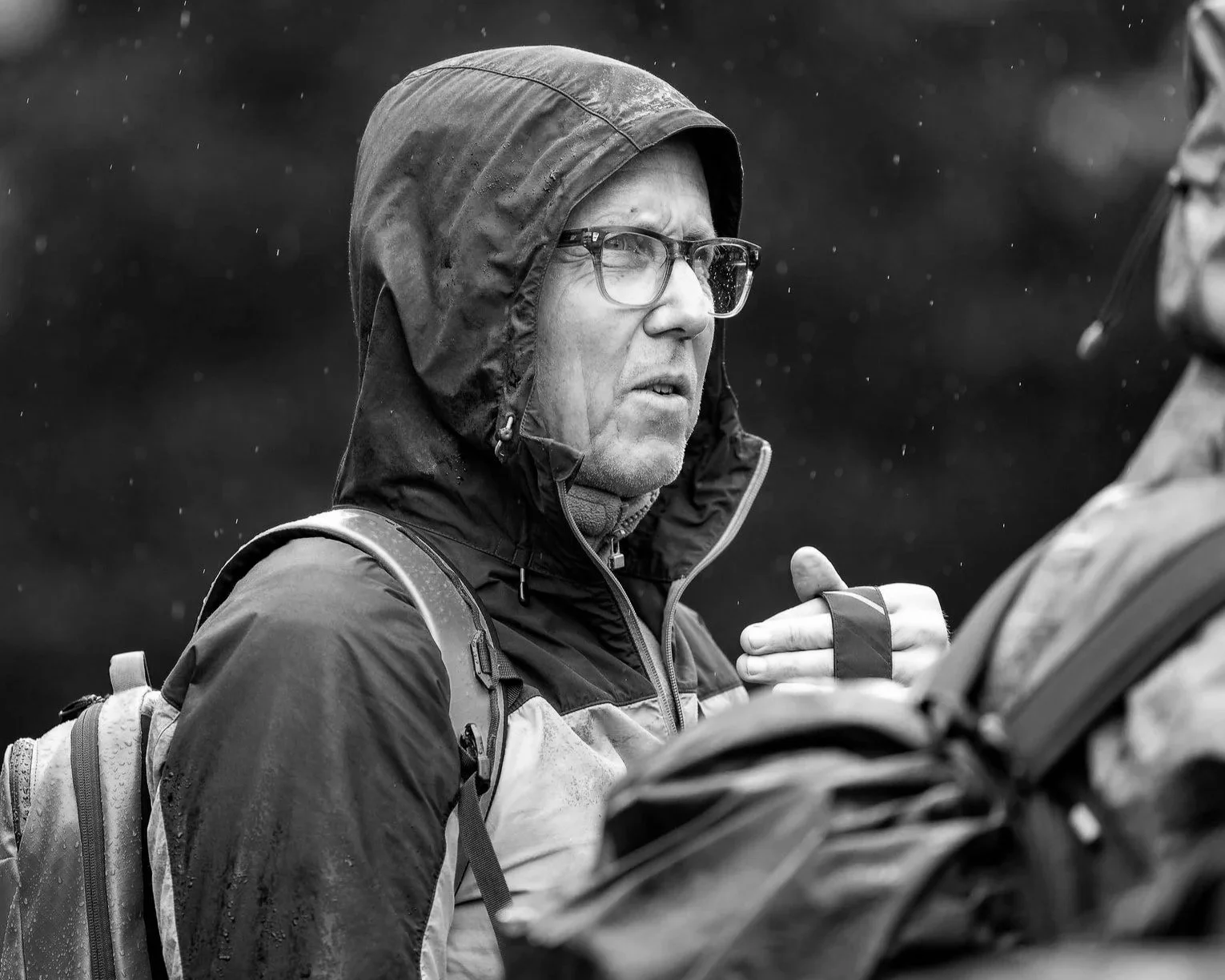 Ein Mann trägt Regenjacke mit Kapuze und Brille, steht im Regen und spricht mit einer Person, die nur teilweise sichtbar ist, während er eine Tasche auf seinem Rücken trägt.