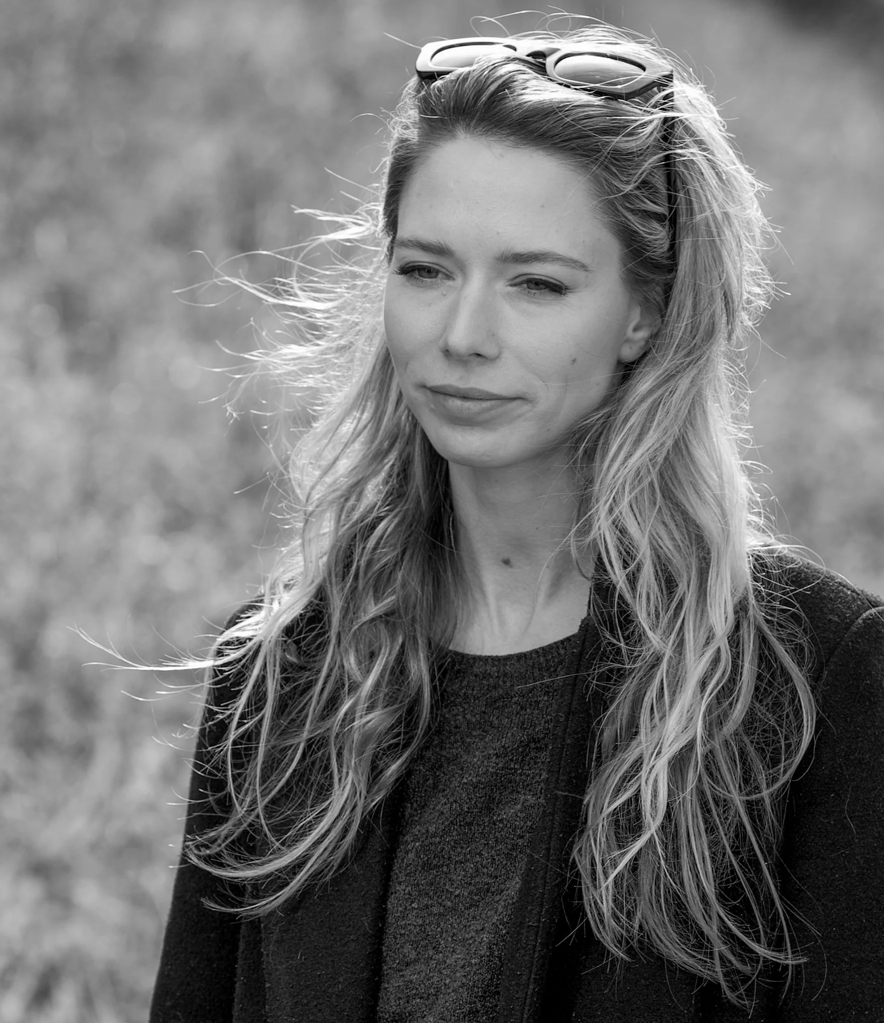 Schwarz-Weiß-Foto einer jungen Frau mit langen, gewellten Haaren, Sonnenbrille auf dem Kopf, trägt eine dunkle Jacke und einen dunklen Pullover, steht im Freien mit verschwommenem Hintergrund.