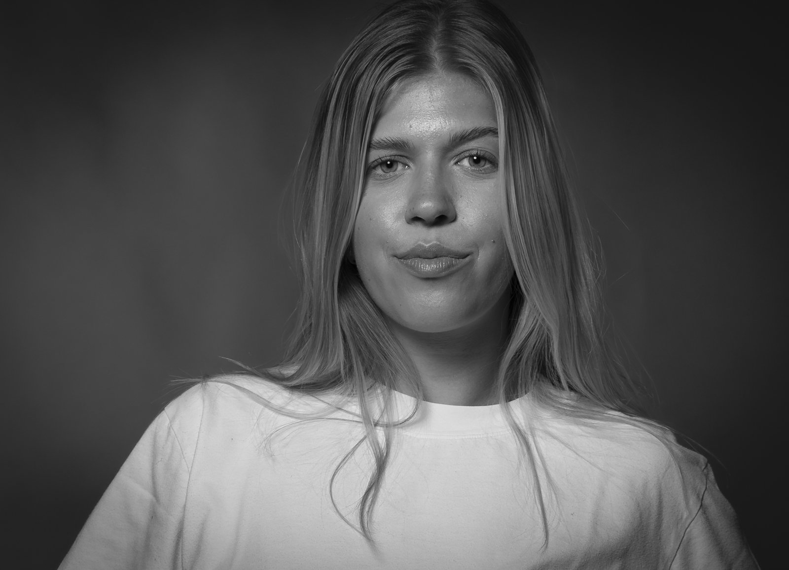Schwarz-weiß Porträt einer jungen Frau mit langem, geradem Haar, trägt ein einfaches weißes T-Shirt, vor dunklem Hintergrund, direkt in die Kamera blickend.