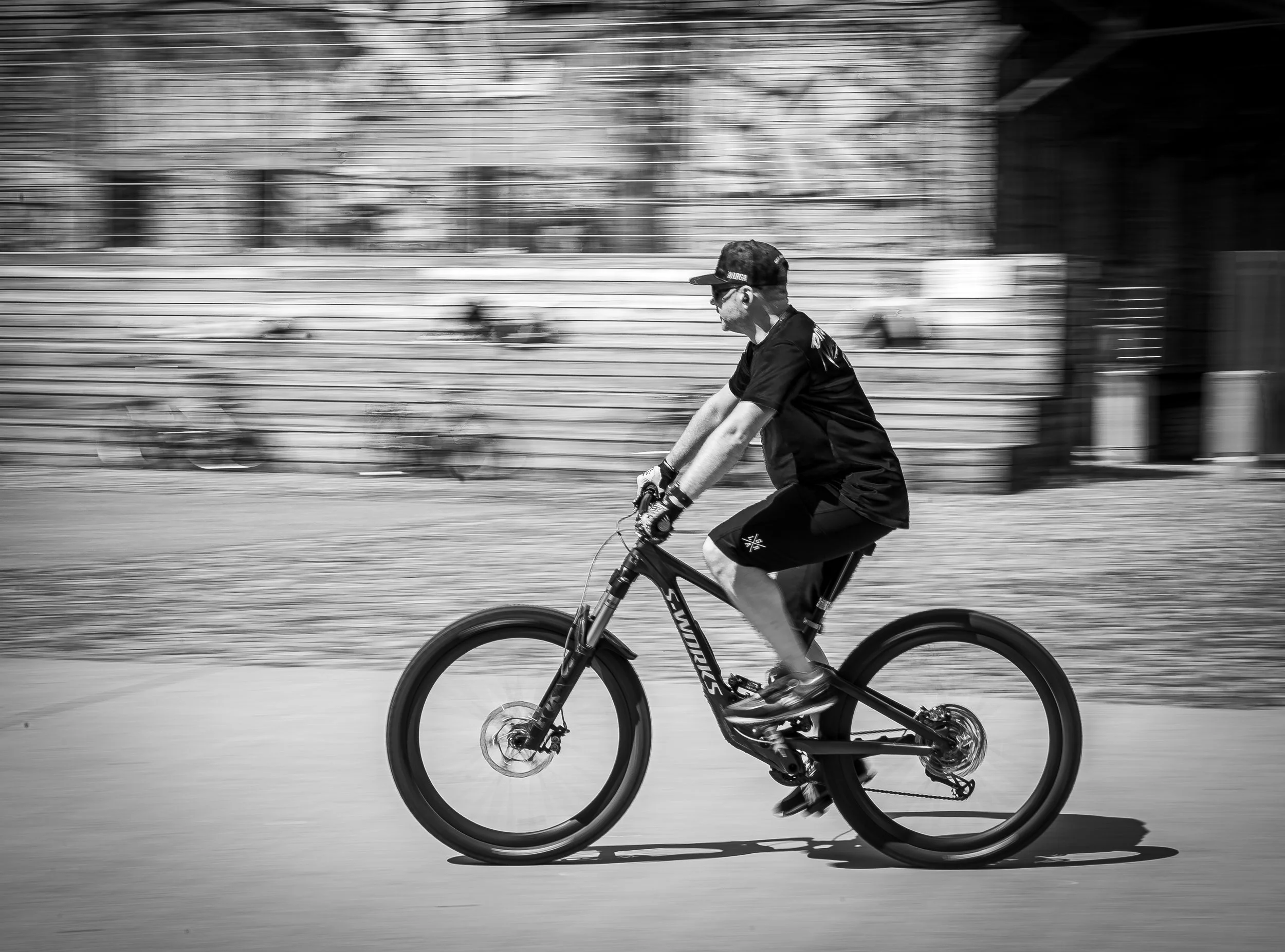 Ein Mann fährt mit einem Mountainbike, schwarzes T-Shirt und Shorts, in Bewegung auf einer offenen Fläche, im Hintergrund sind Bänke und Bäume, im Schwarz-Weiß-Stil.