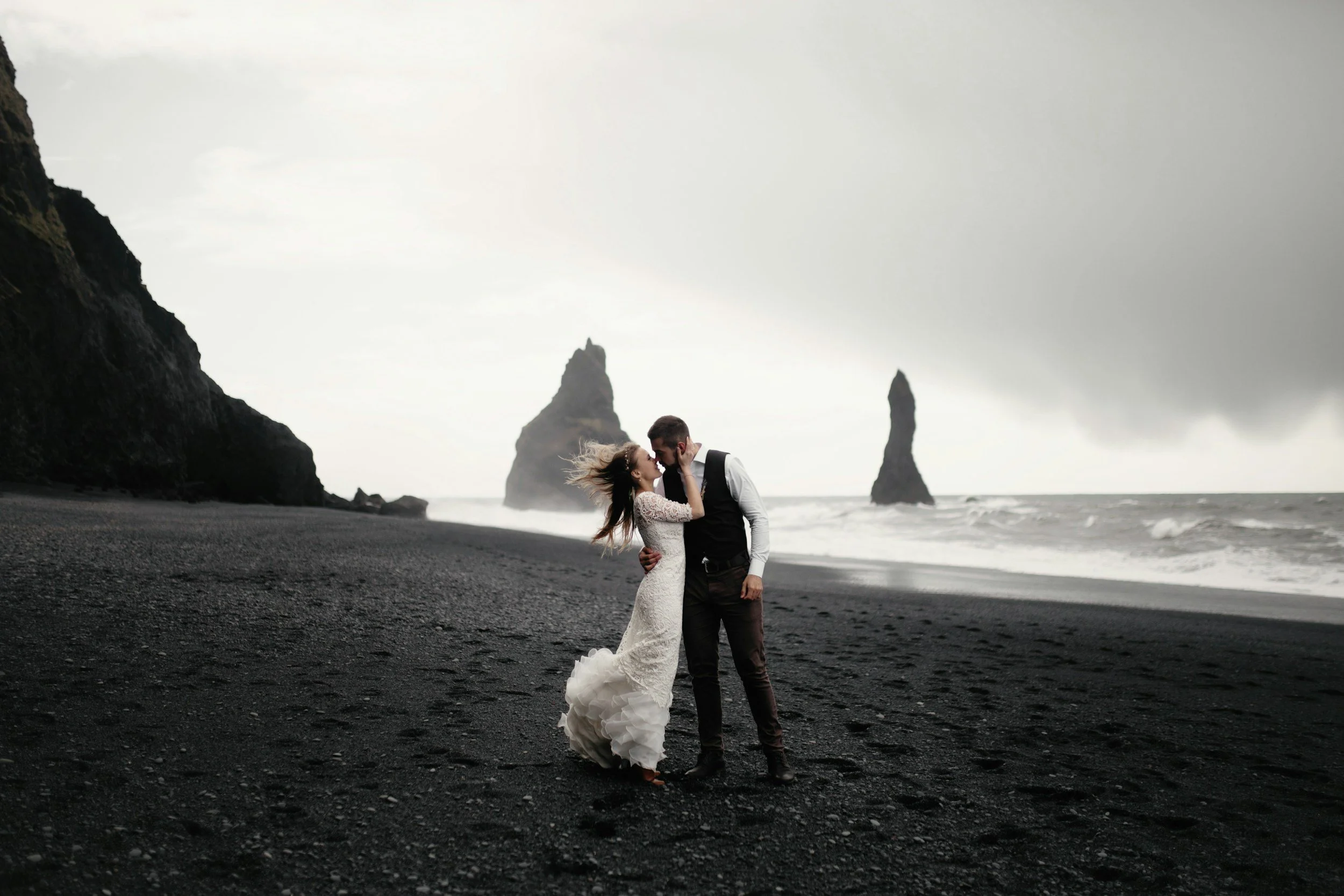 A couple in wedding attire standing on a black sand beach, embracing and touching foreheads, with tall rock formations in the background under a cloudy sky.