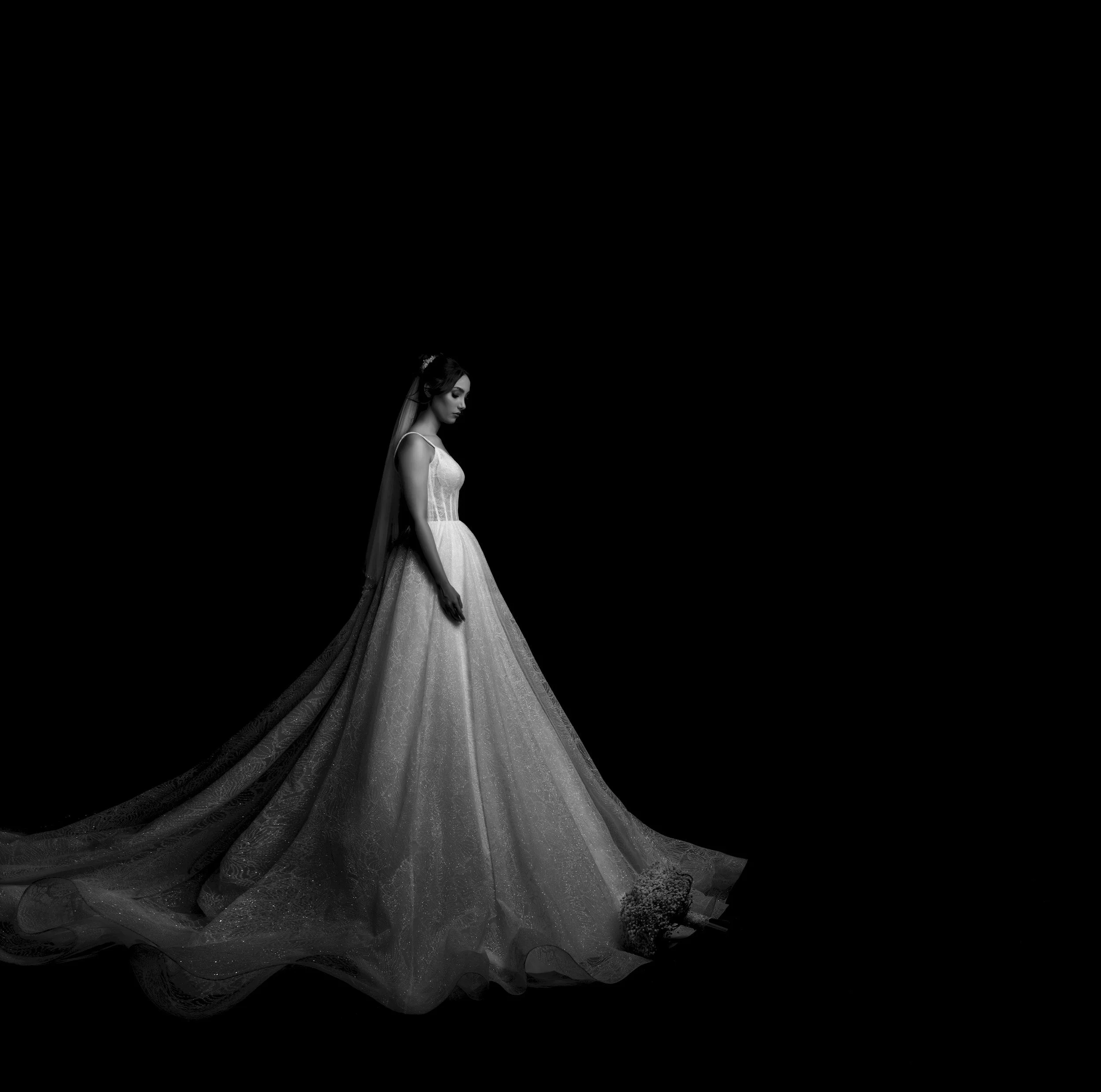 Black and white photo of a woman in a wedding dress with a long train, standing against a dark background.