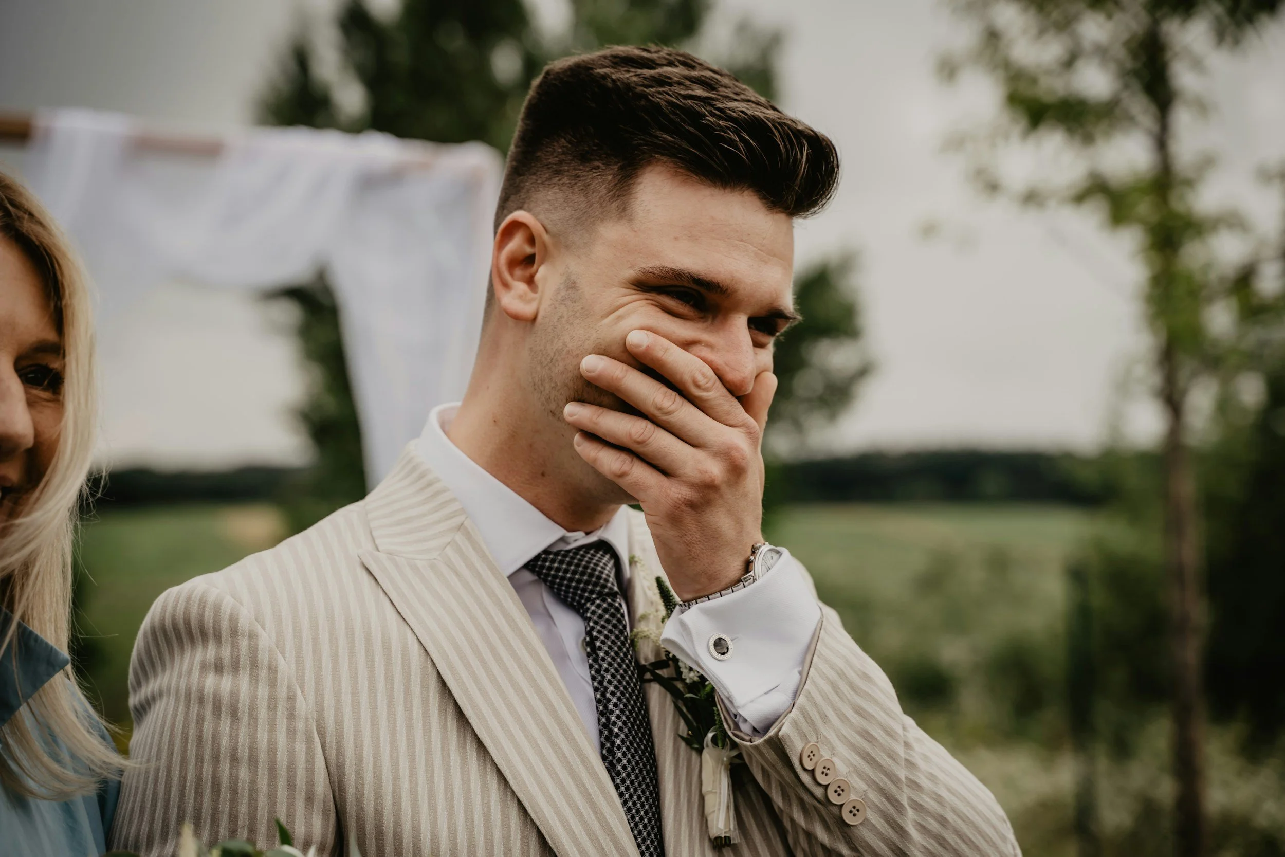 A man in a beige pinstripe suit covering his mouth and smiling at an outdoor event, with a woman partially visible on the left side, in front of a background of trees and a white cloth hanging on a line.