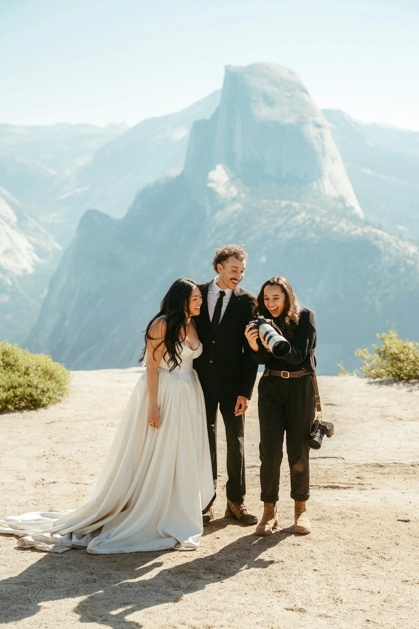 A bride and groom with a female photographer, standing outdoors on a sandy surface, looking at a camera held by the photographer, with a mountainous landscape featuring a tall, rounded rock formation in the background.