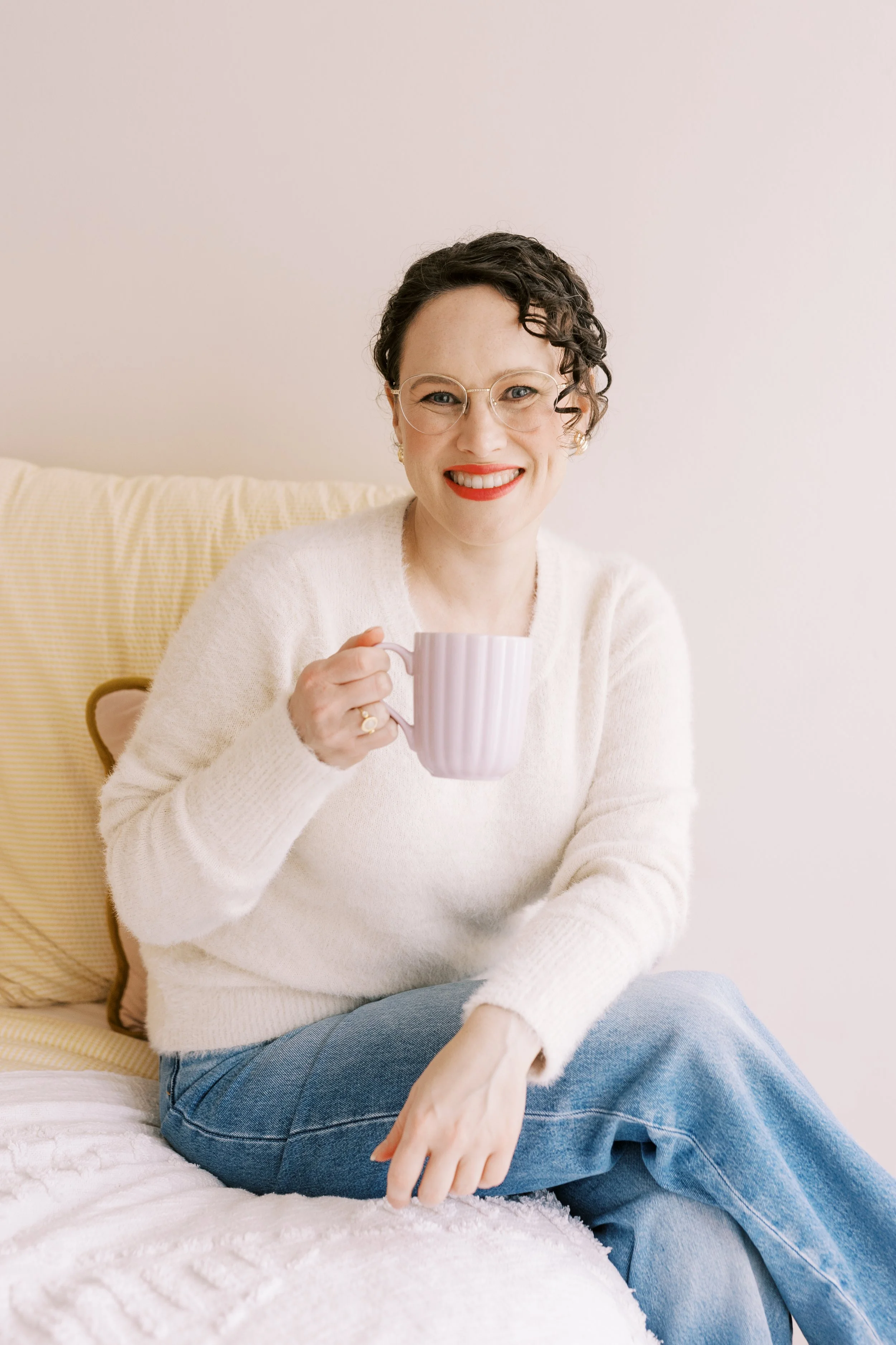 A woman with short curly hair, glasses, and red lipstick, smiling and holding a pink mug, sitting on a bed with a light-colored blanket and yellow pillows, against a plain light wall.