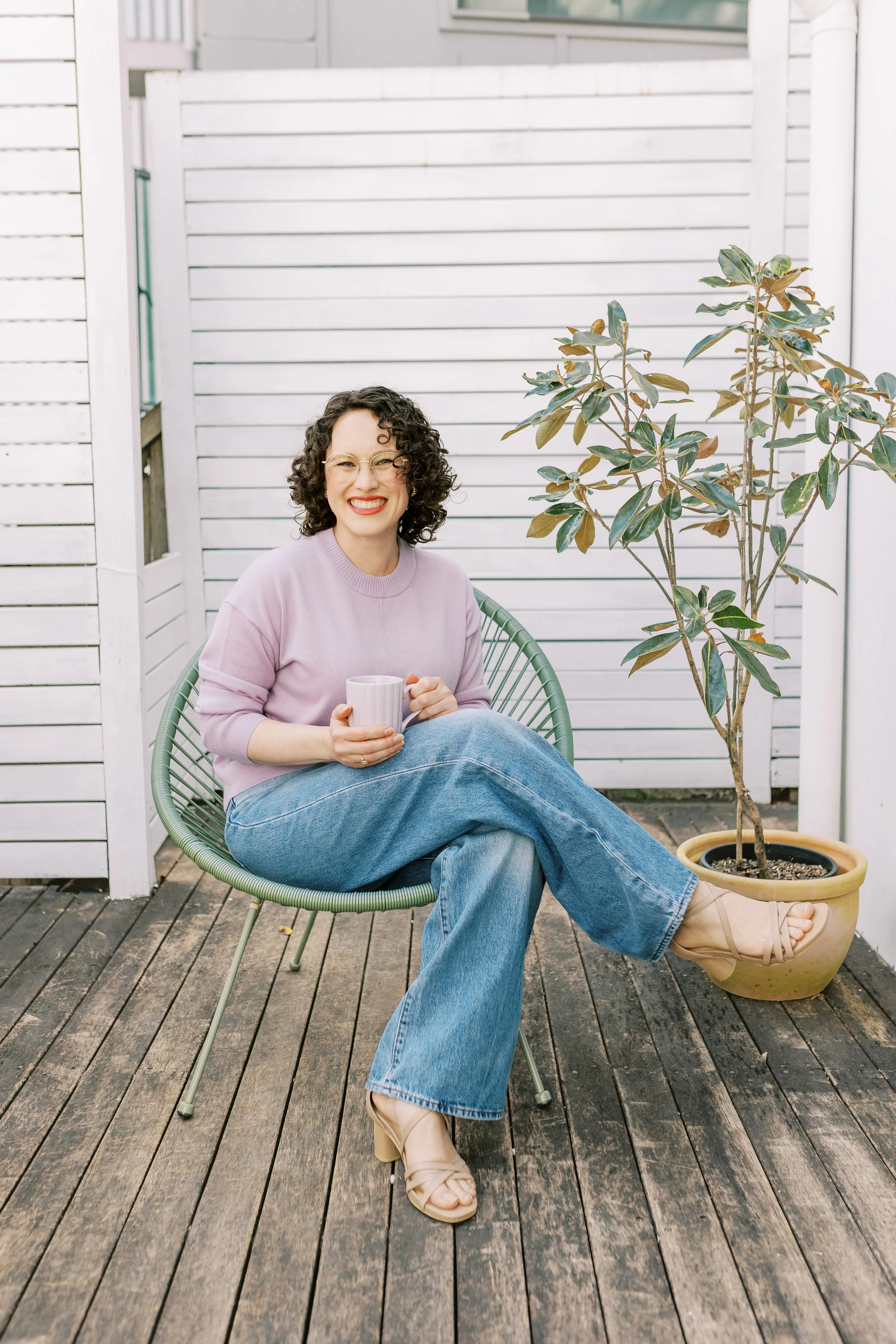 A woman with curly hair, glasses, and red lipstick sitting outside on a wooden deck, smiling while holding a mug with a large potted plant nearby, against a white wooden fence background.
