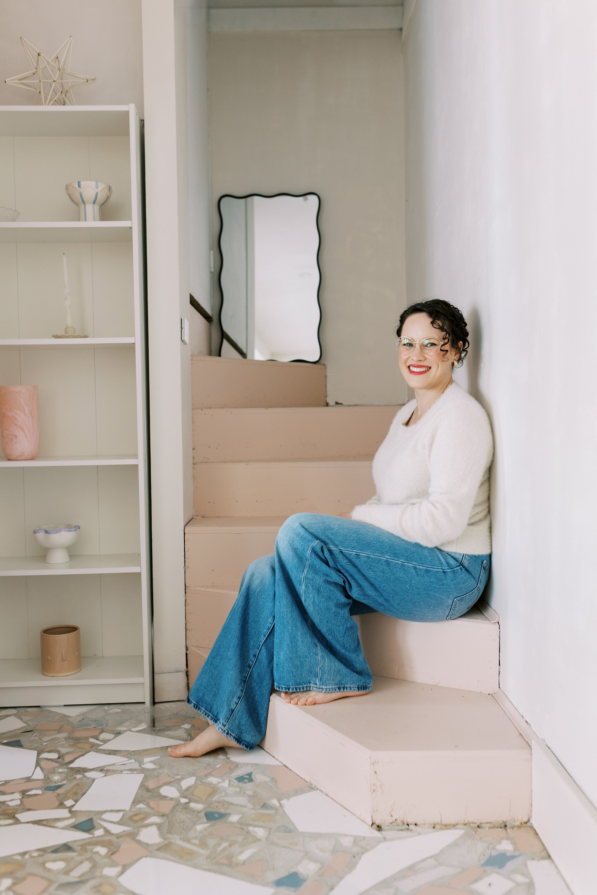 A woman with glasses, curly hair, and red lipstick sitting on stairs in a bright room, wearing a white sweater and blue jeans, smiling at the camera.