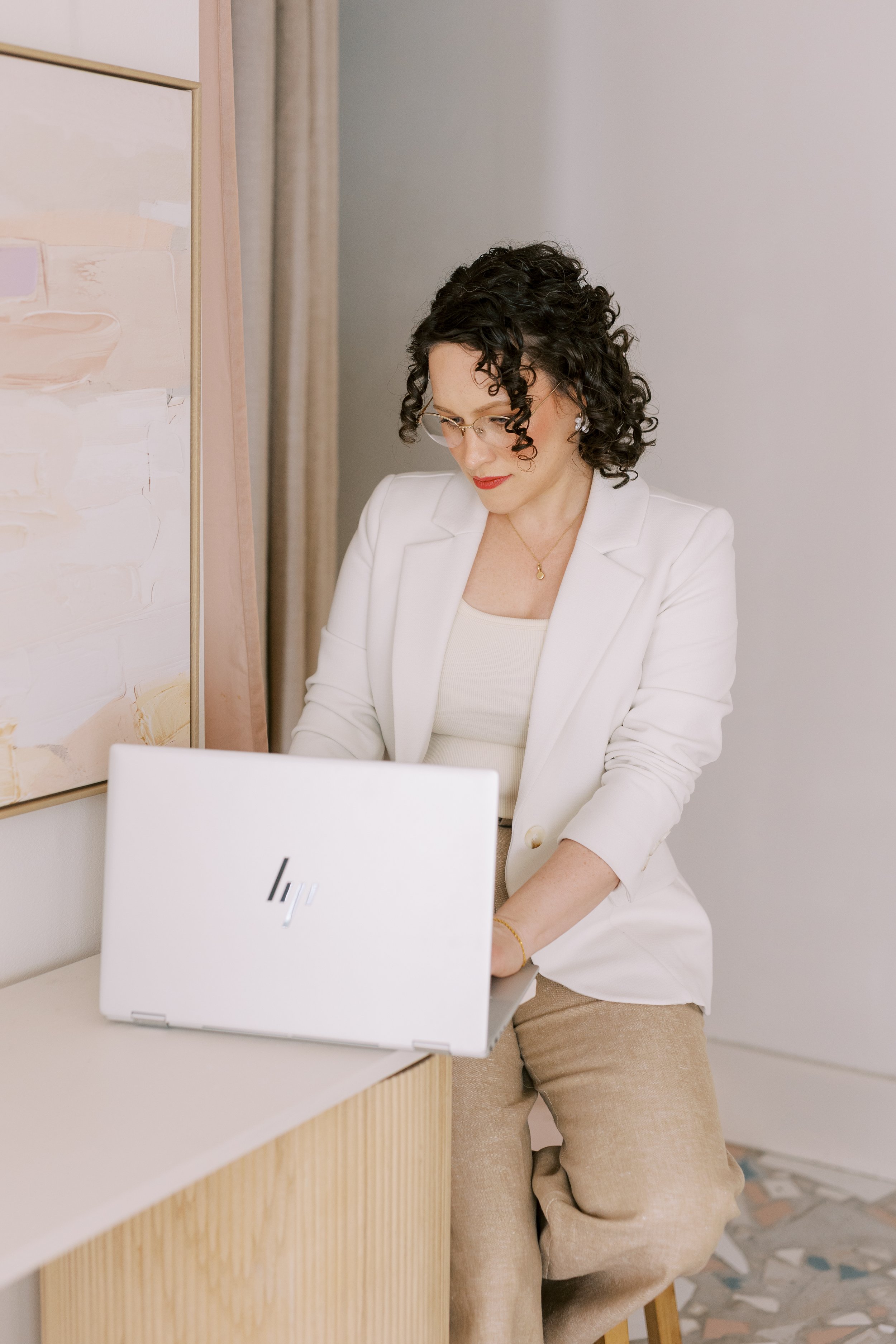 Woman with curly dark hair, glasses, and red lipstick in a white blazer working on a silver HP laptop in a bright room.