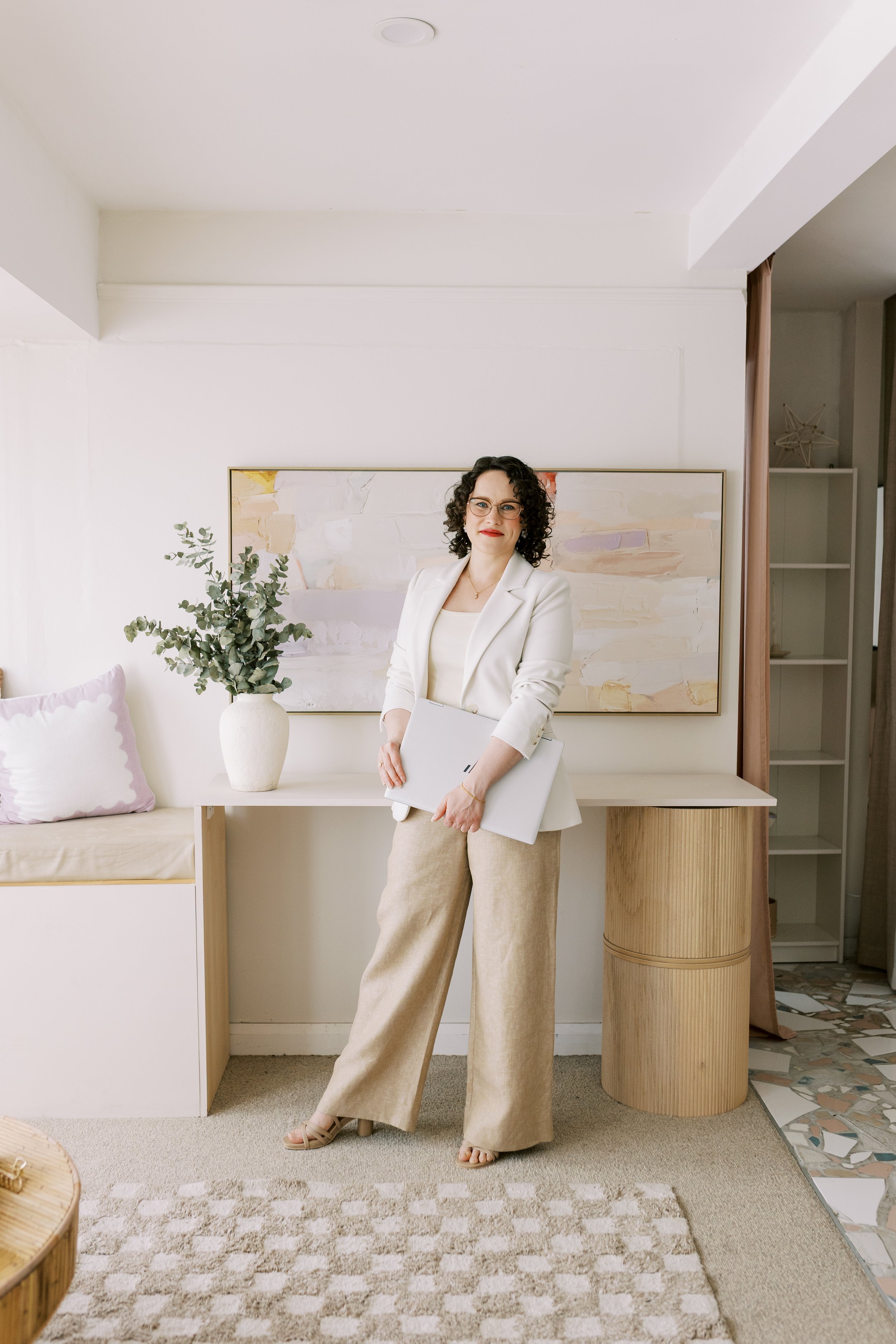 A woman with curly dark hair and glasses, wearing a white blazer and beige pants, stands in a modern, minimalist living room holding a silver laptop.