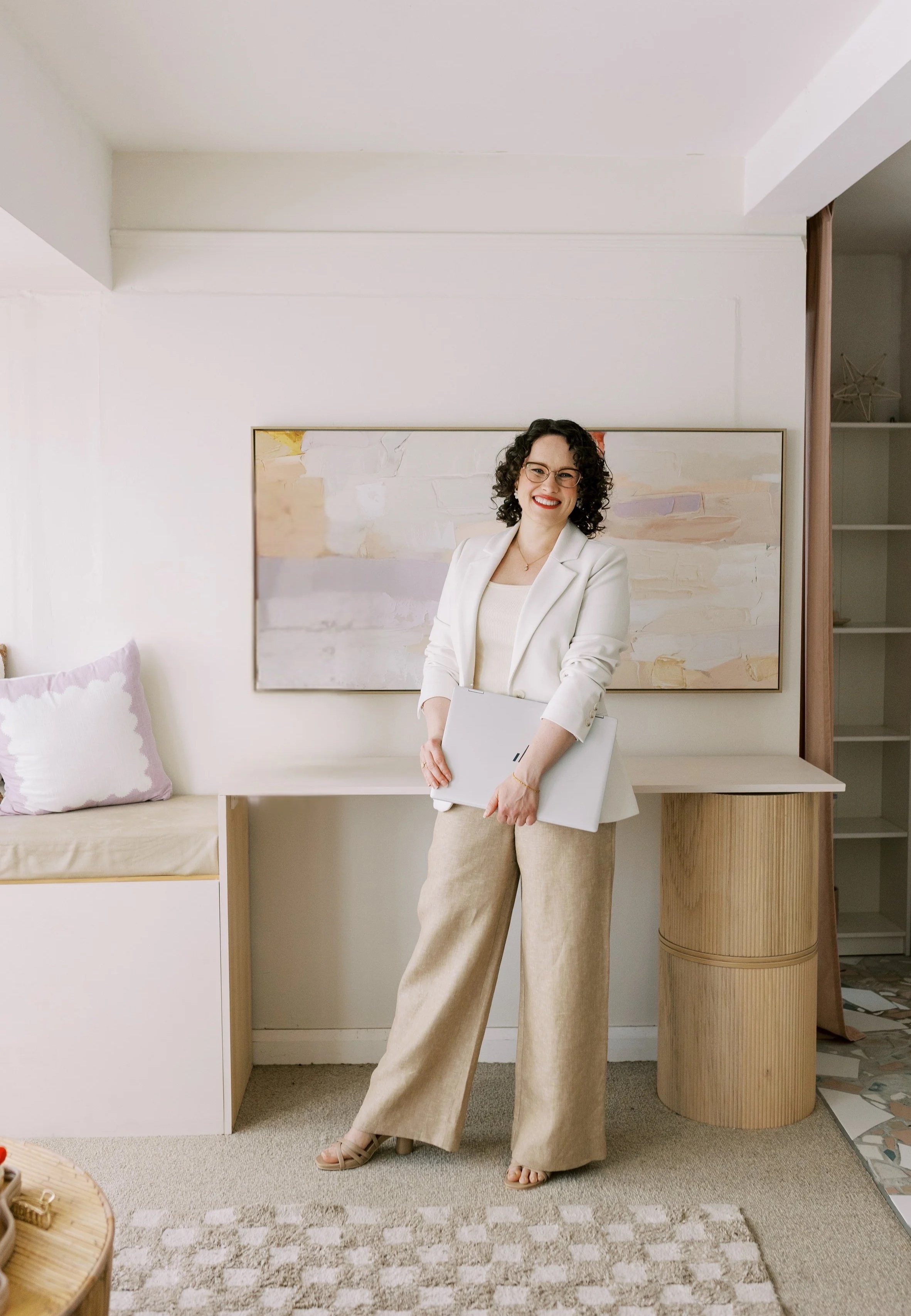 A woman in a white blazer and beige pants holding a laptop, standing in a modern, minimalistic room with abstract artwork and decorative pillows.