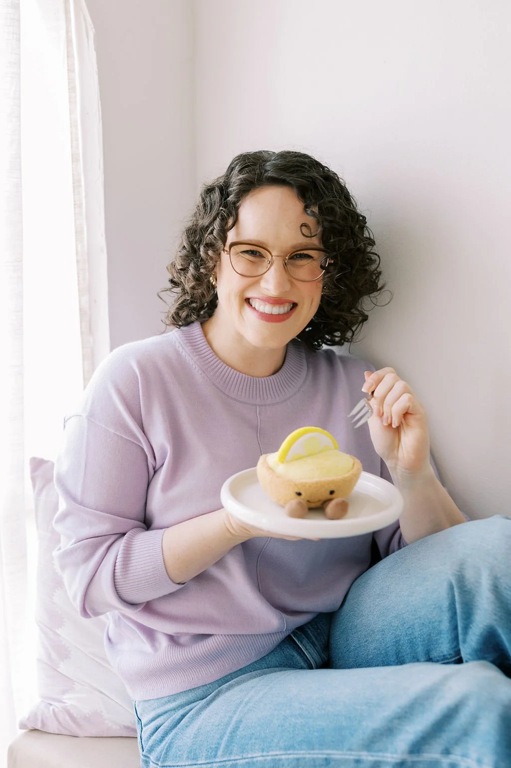 Woman with curly hair and glasses smiling while holding a plate with a lemon-themed pastry, sitting near a window.