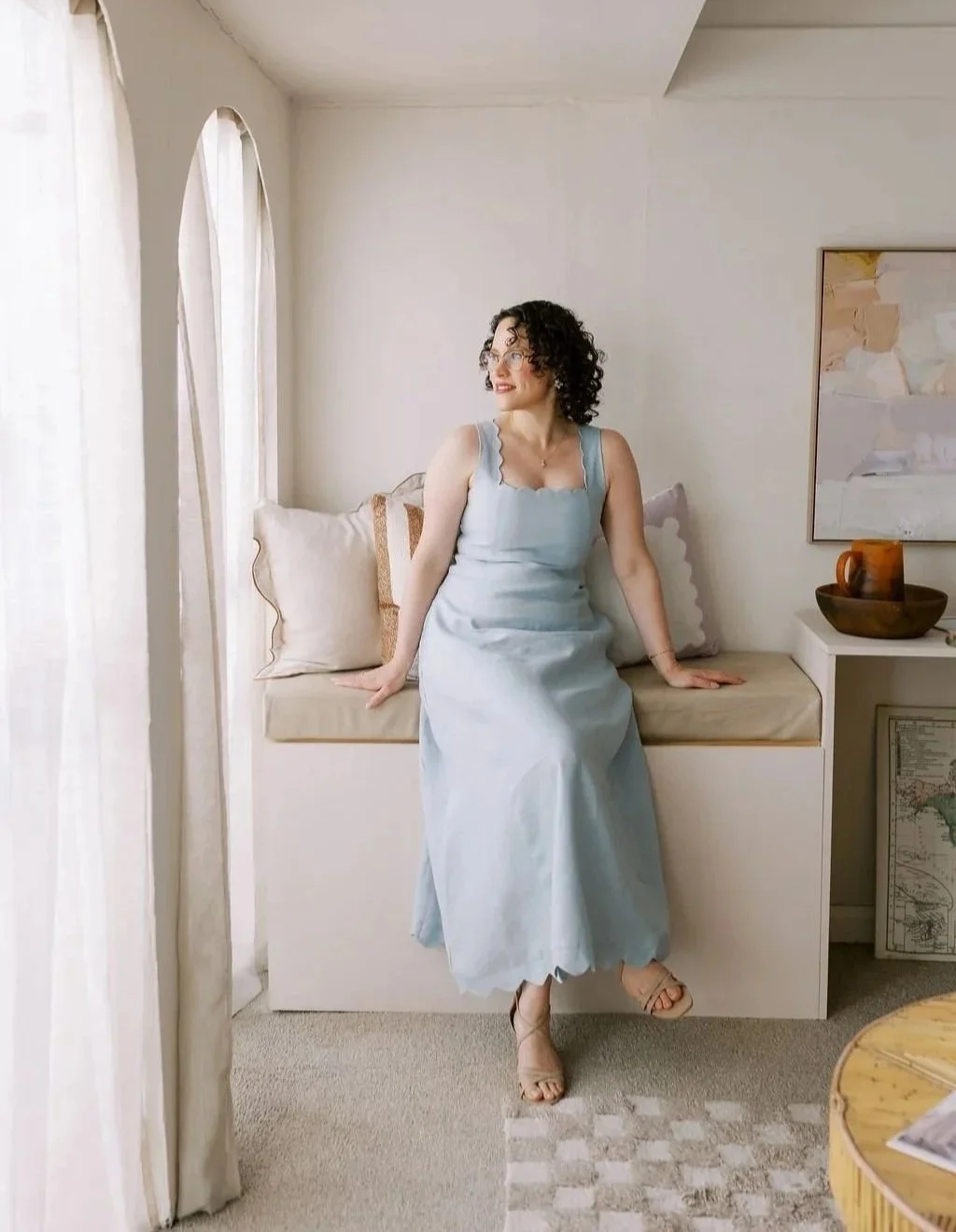 A woman with curly hair and glasses is sitting on a built-in window seat in a bright room, wearing a light blue dress and heels, looking out to the side.